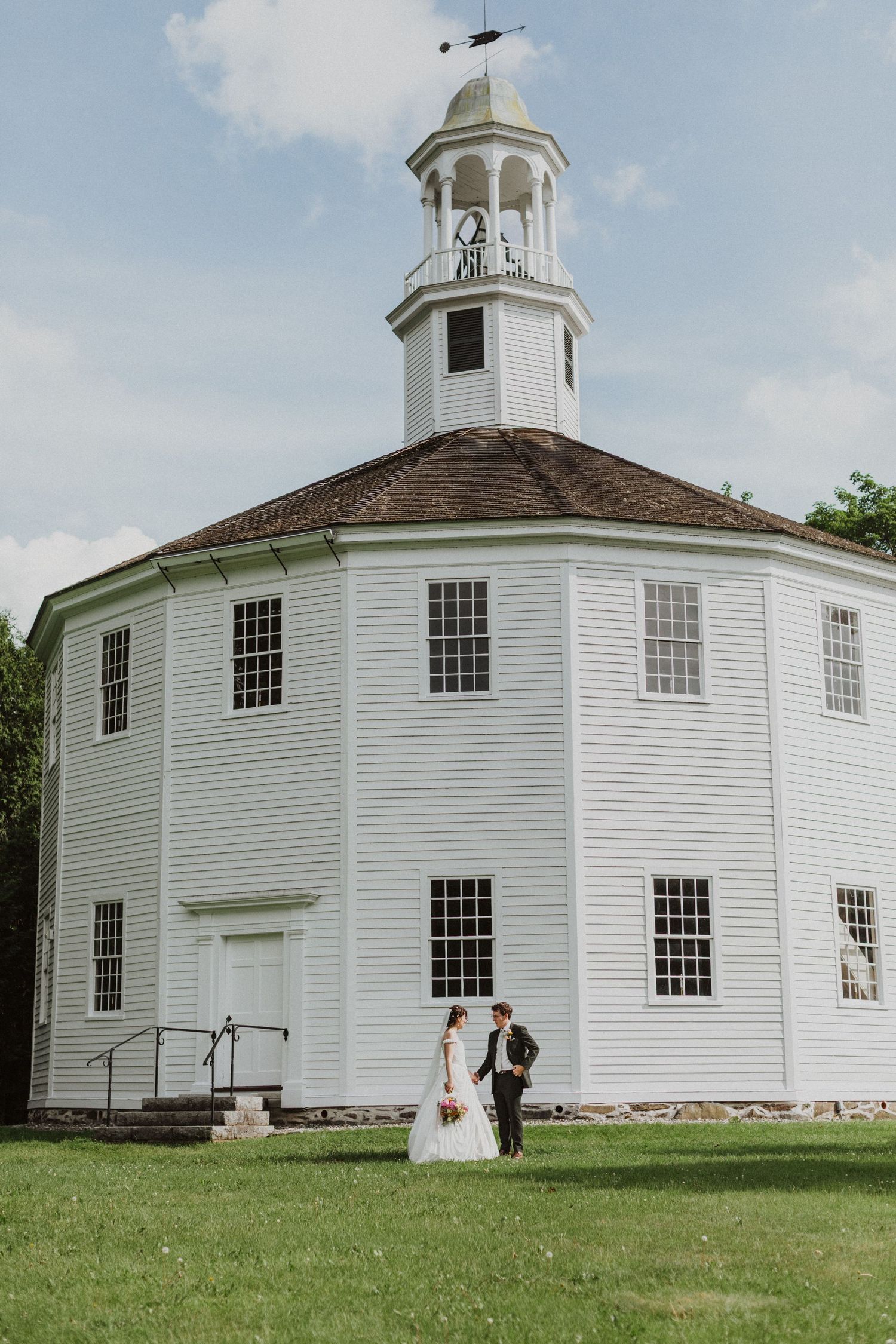 Bride and groom posing in front of a historic white octagonal church on a sunny wedding day.