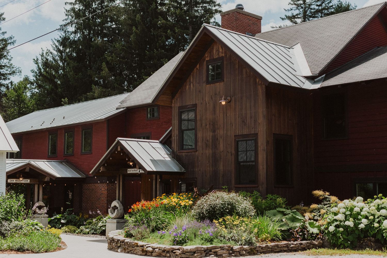 Rustic red barn wedding venue with metal roof, lush gardens, and tall pine trees in a Vermont countryside setting.