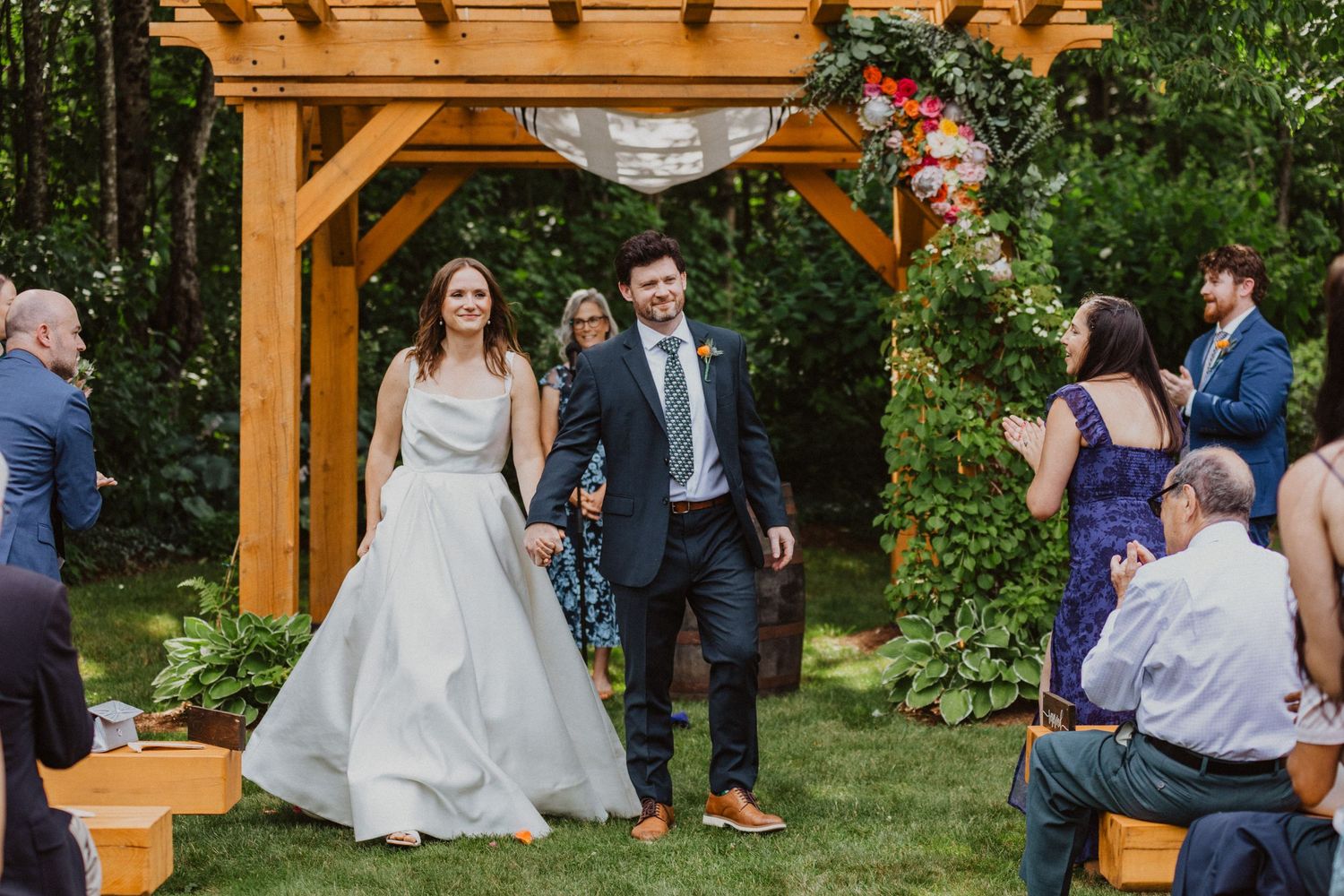 A joyful newlywed couple walks hand-in-hand under a wooden arbor surrounded by guests at an outdoor wedding.