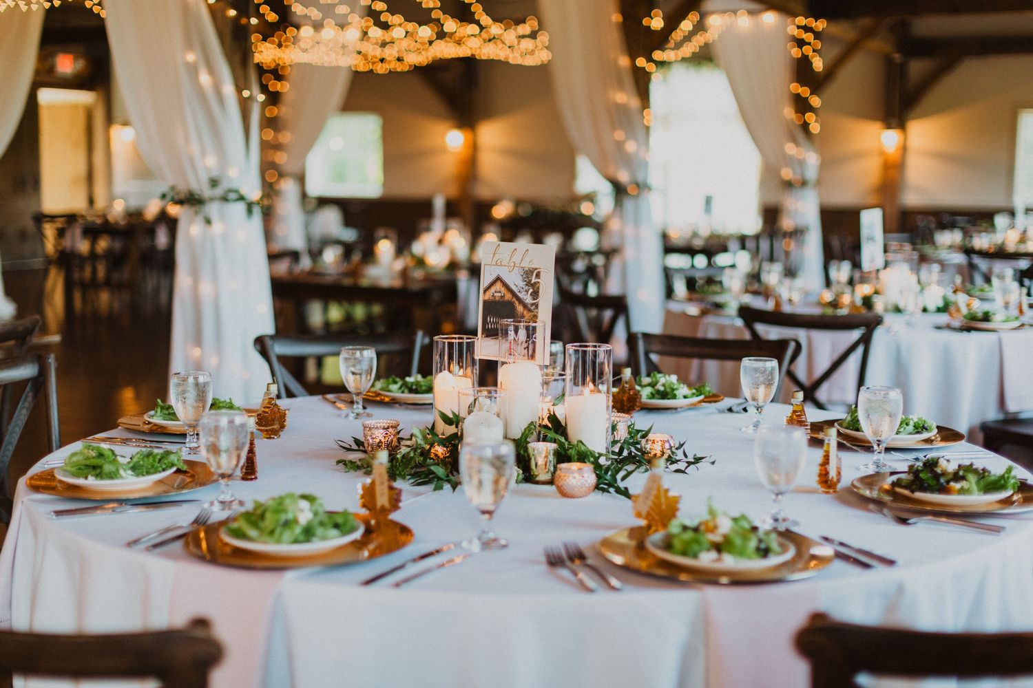 Elegant wedding reception table setting with white linens, candles, and greenery centerpieces.