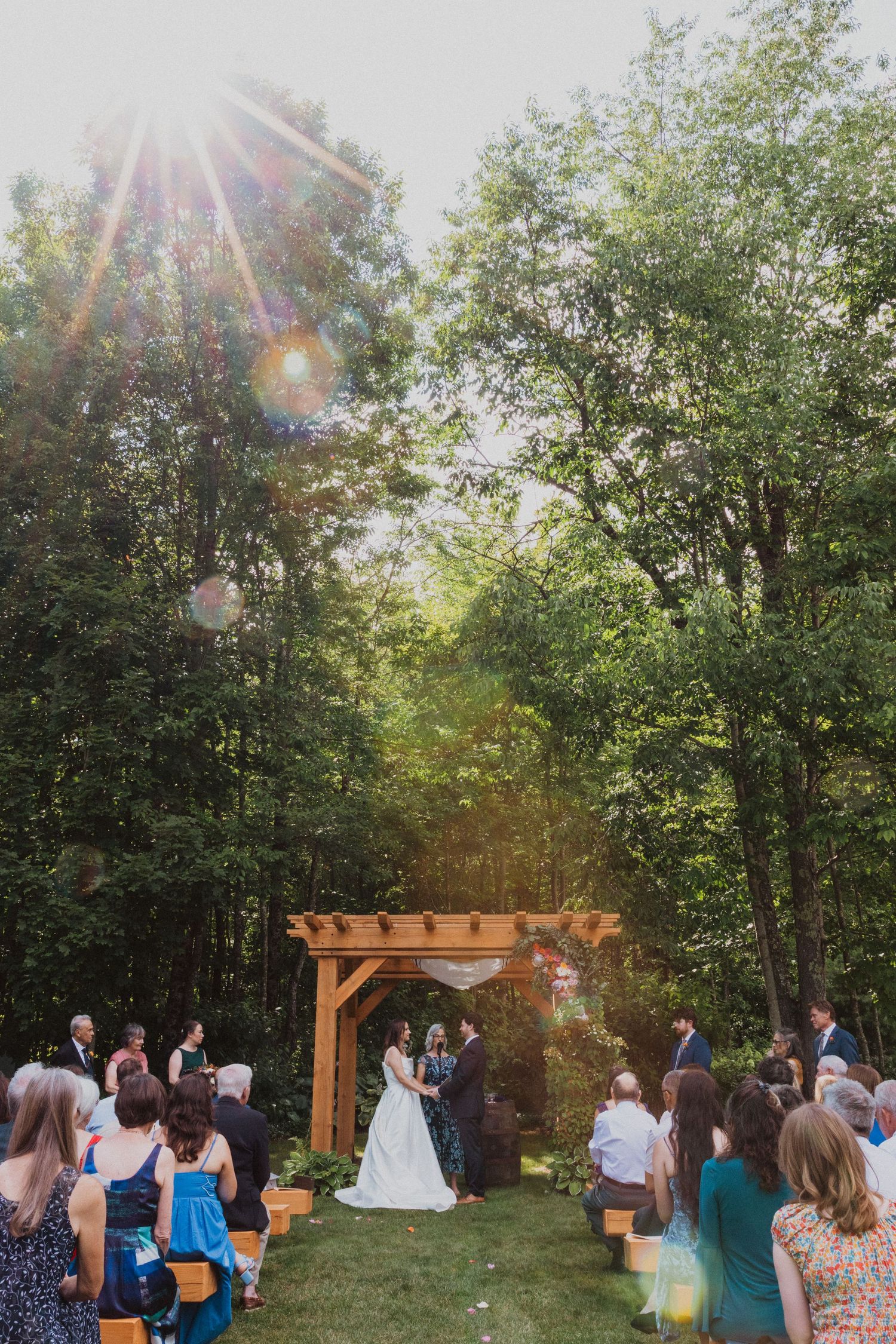 Outdoor wedding ceremony under wooden pergola surrounded by tall trees with sunlight streaming through.