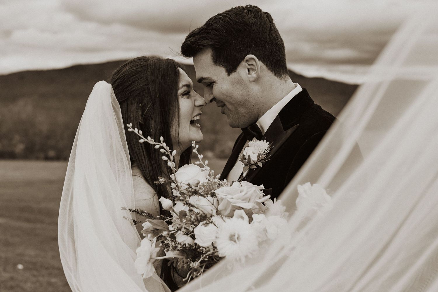 Bride and groom sharing intimate moment wrapped in veil with floral bouquet in black and white photo.