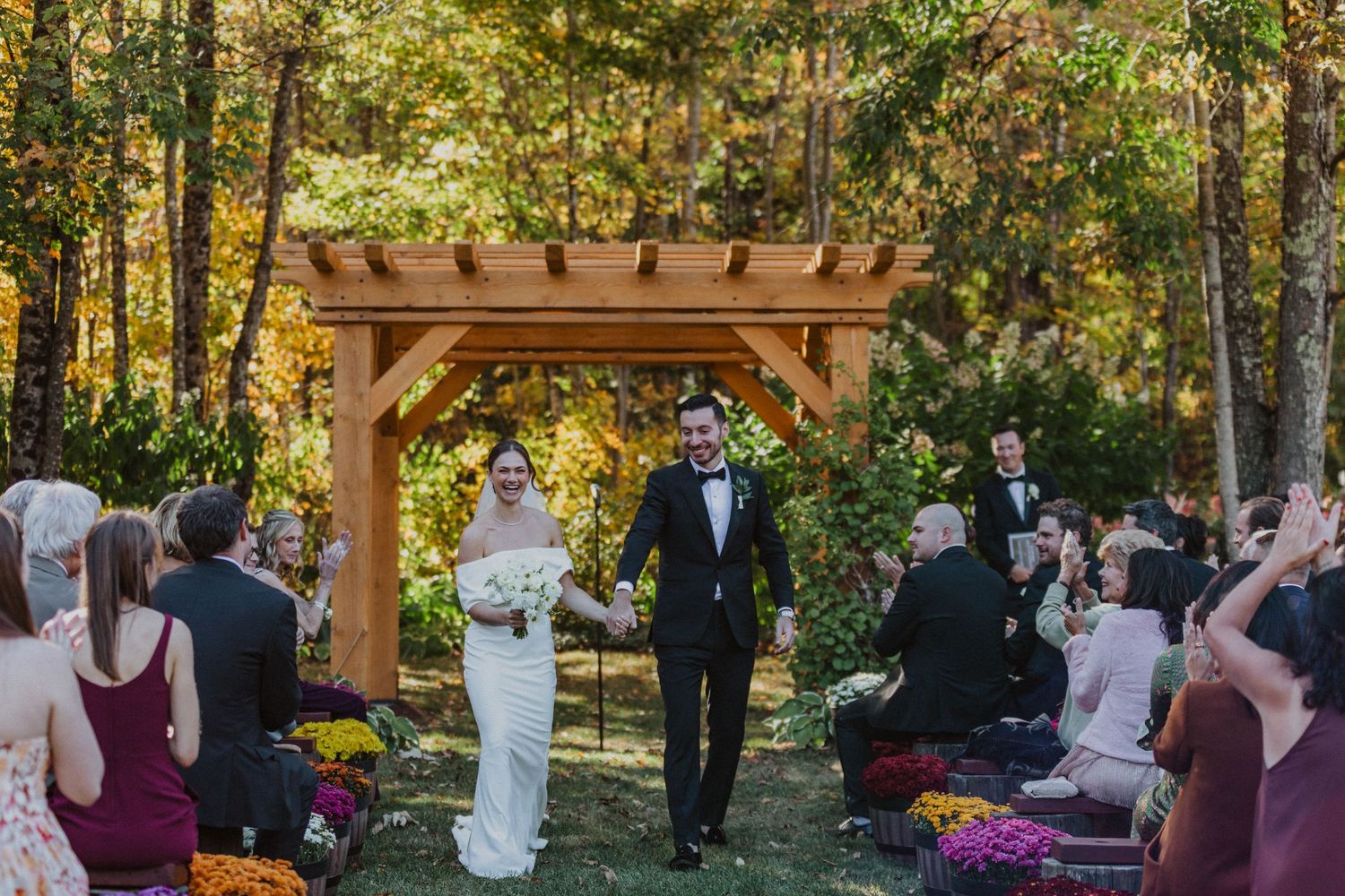 Bride and groom walk down aisle at outdoor fall wedding under wooden pergola arch.