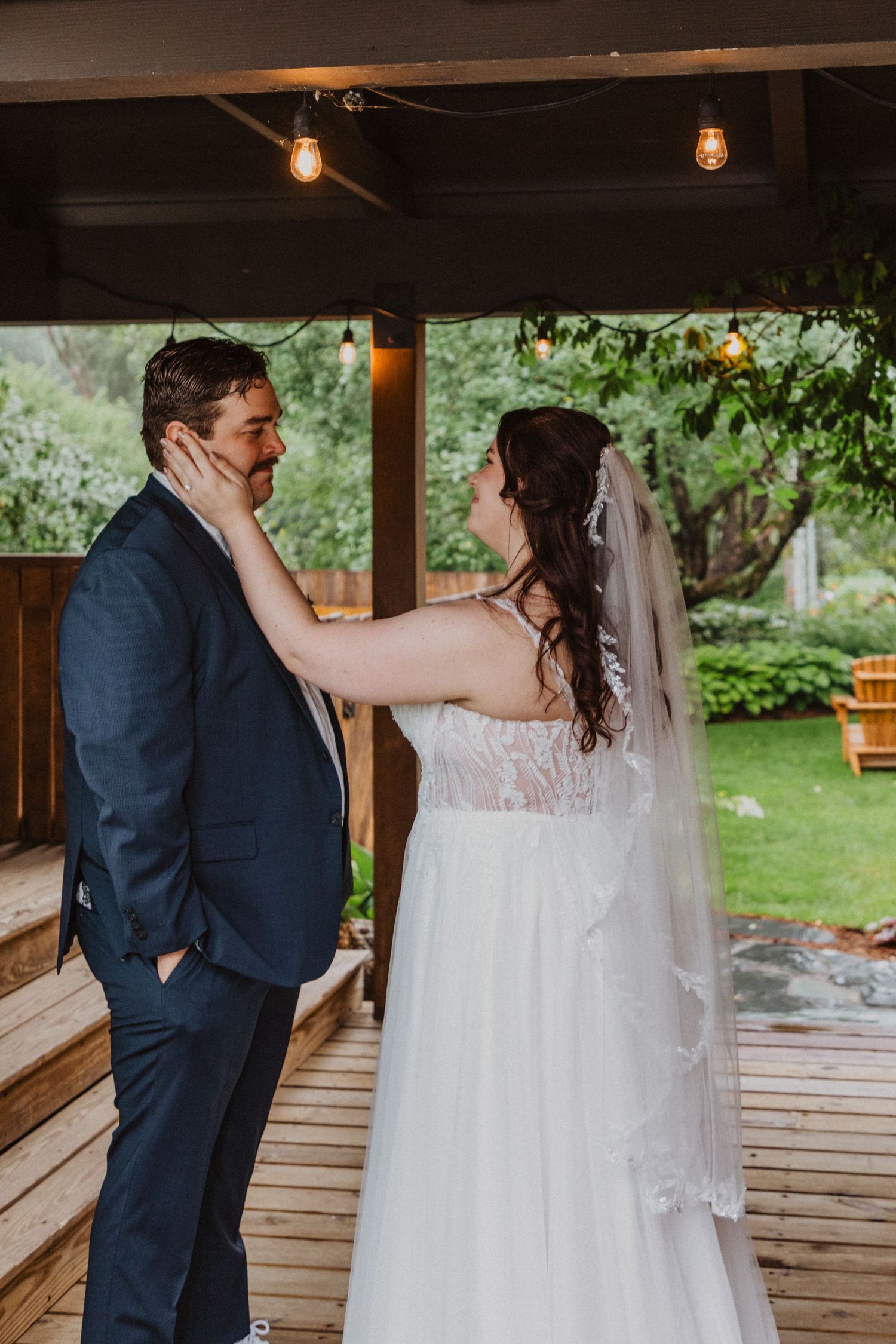 Bride tenderly holds groom's face on rustic wooden deck with string lights at outdoor wedding venue.