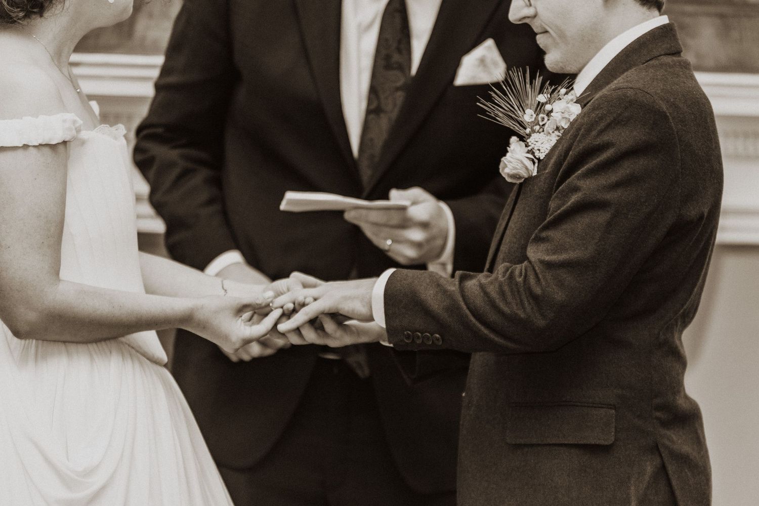 Groom placing wedding ring on brides hand during ceremony in sepia toned photo with officiant holding book.