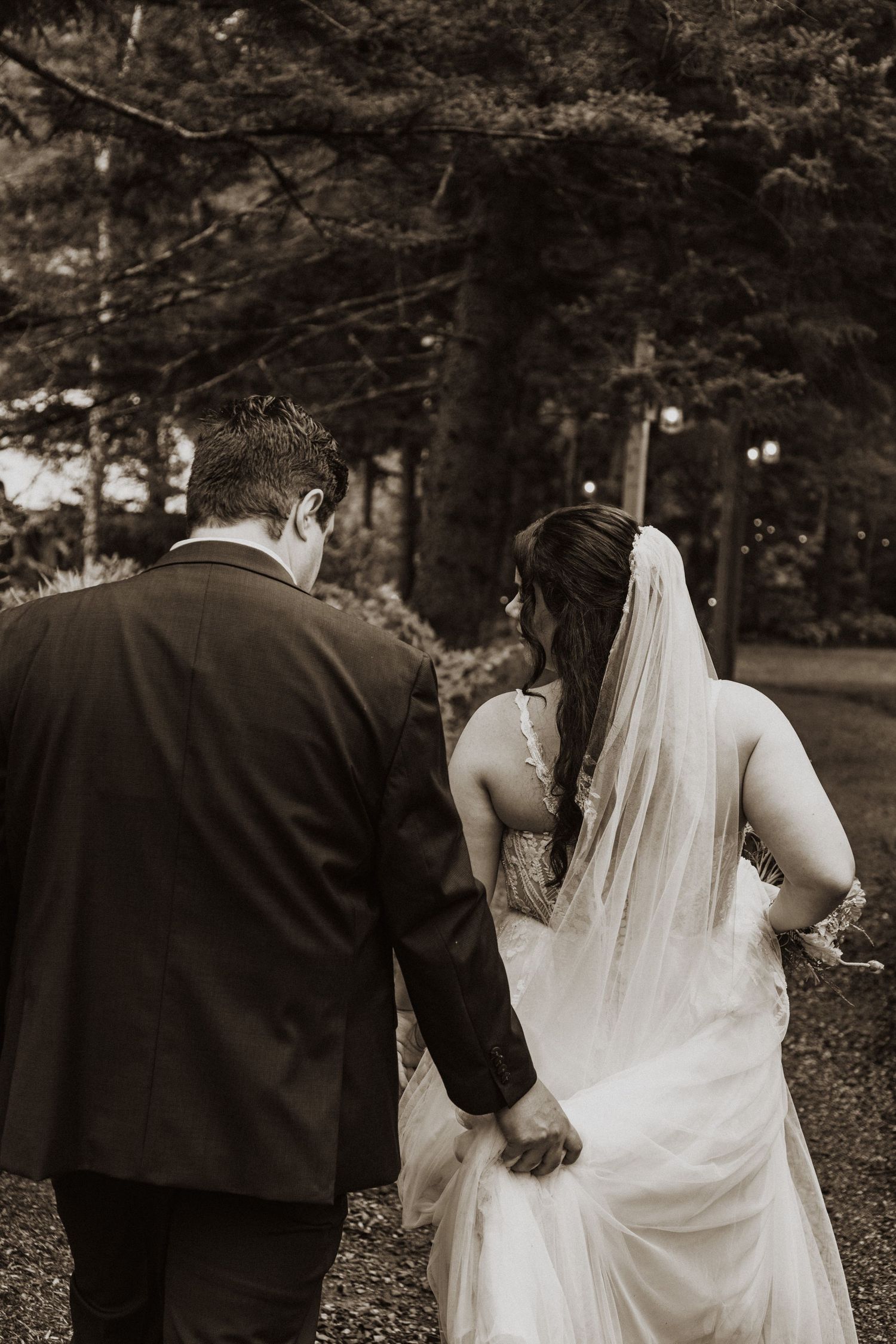 Bride and groom walk hand in hand through lush forest in romantic black and white wedding photo.