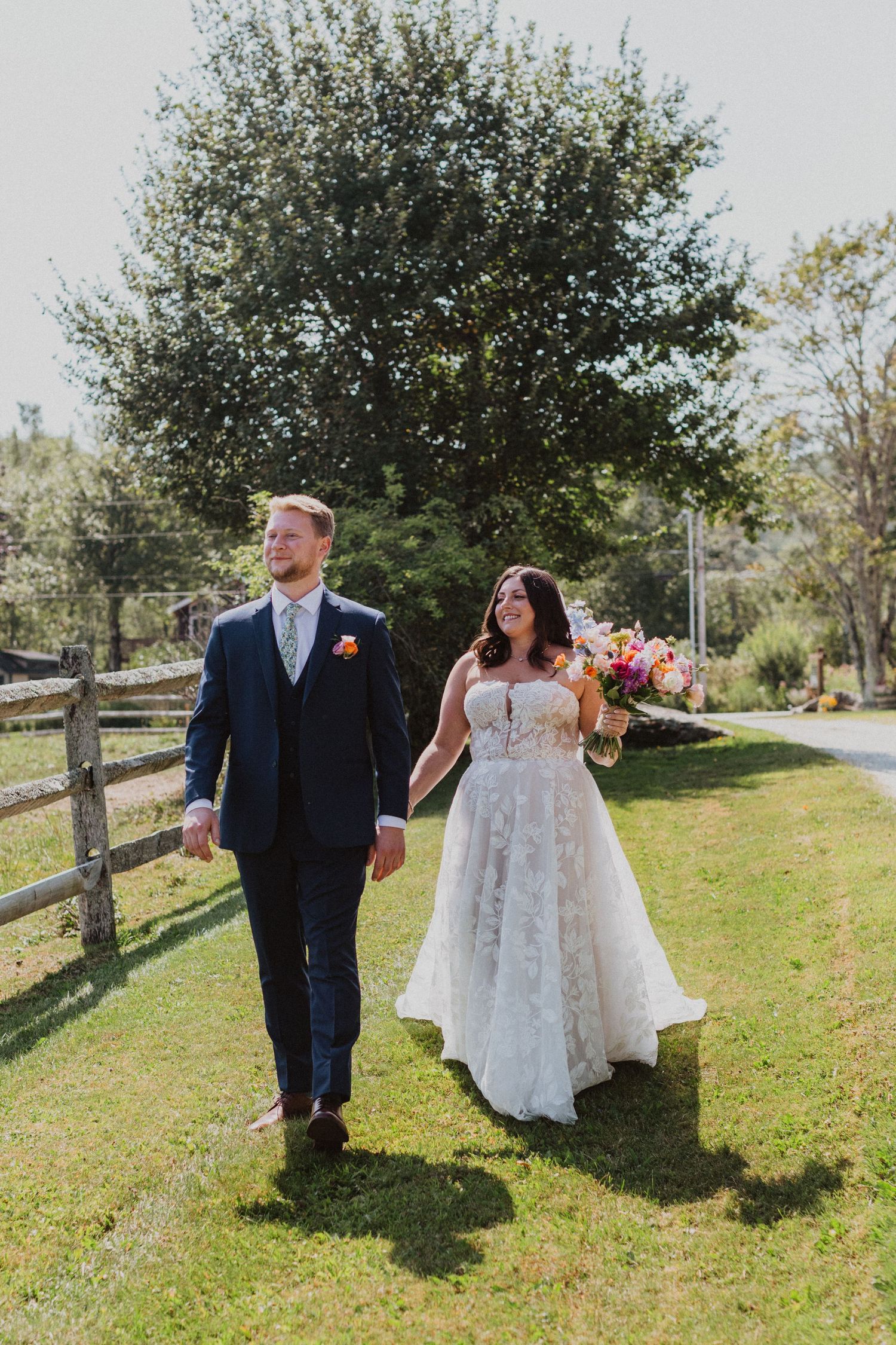 Bride in floral lace gown and groom in navy suit walking hand in hand along wooden fence.