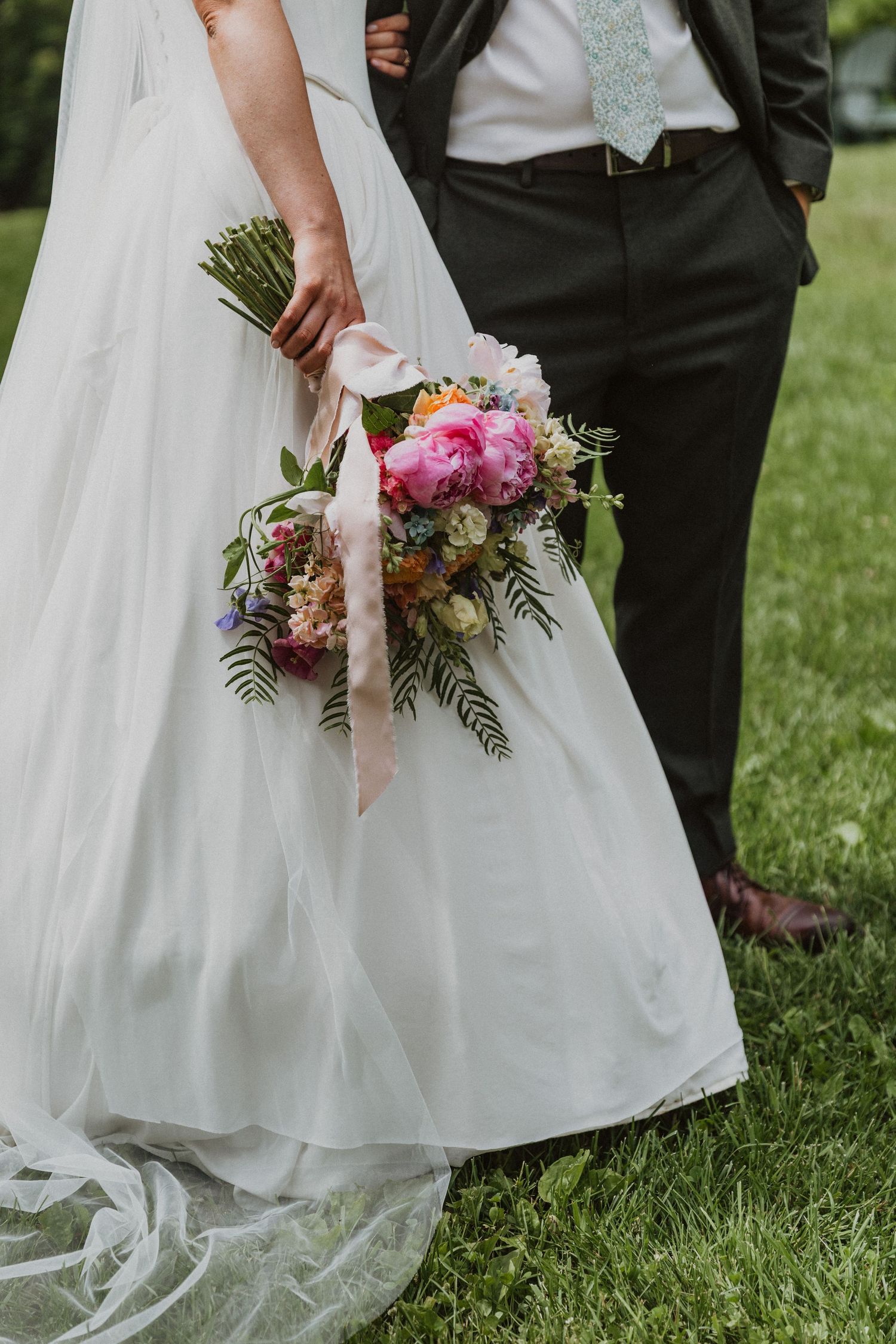 Bride in white gown holding colorful bridal bouquet with pink peonies and ribbons beside groom in dark suit outdoors.