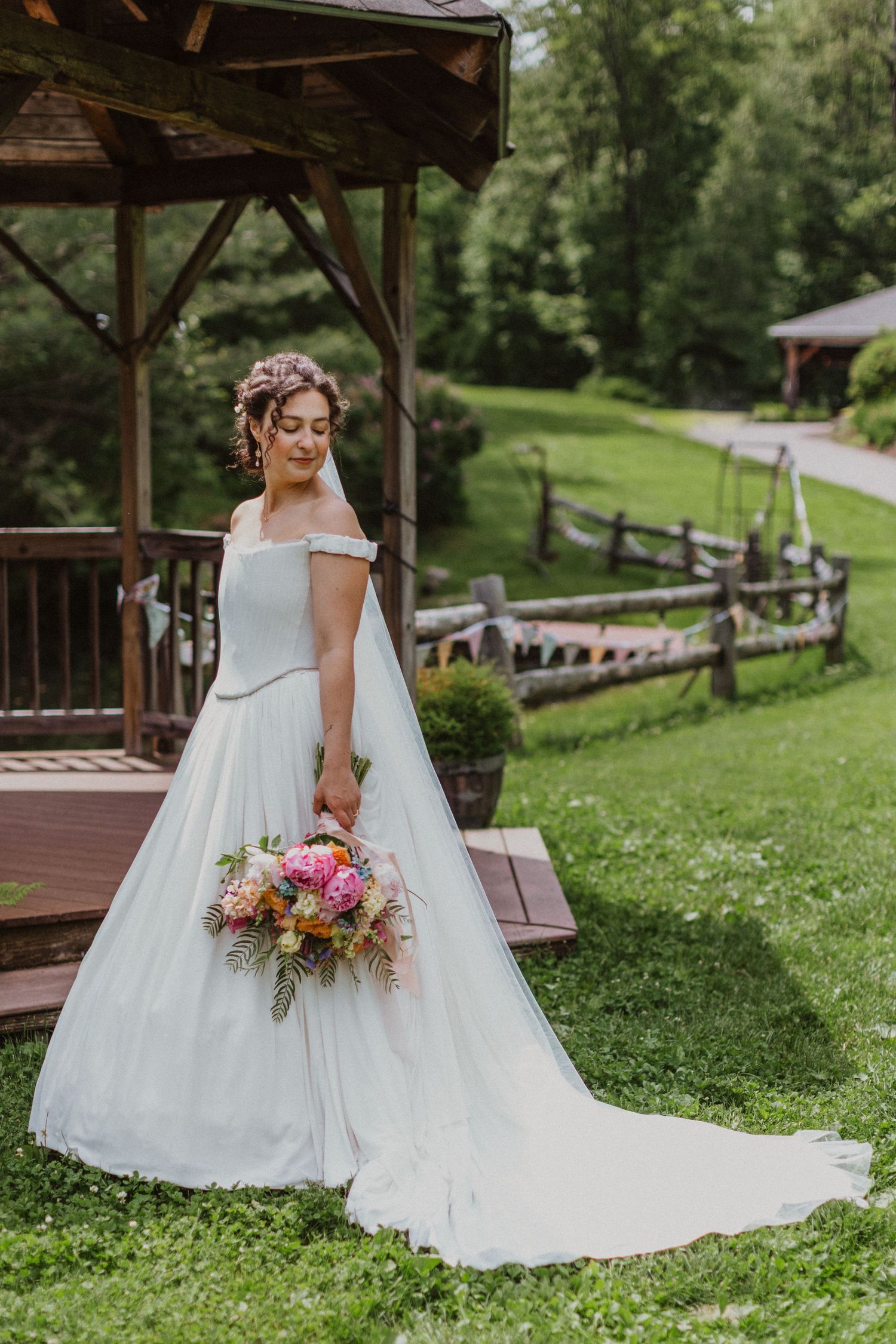 Bride in off-shoulder white ball gown with long train holding colorful bouquet near rustic wooden gazebo in green garden.
