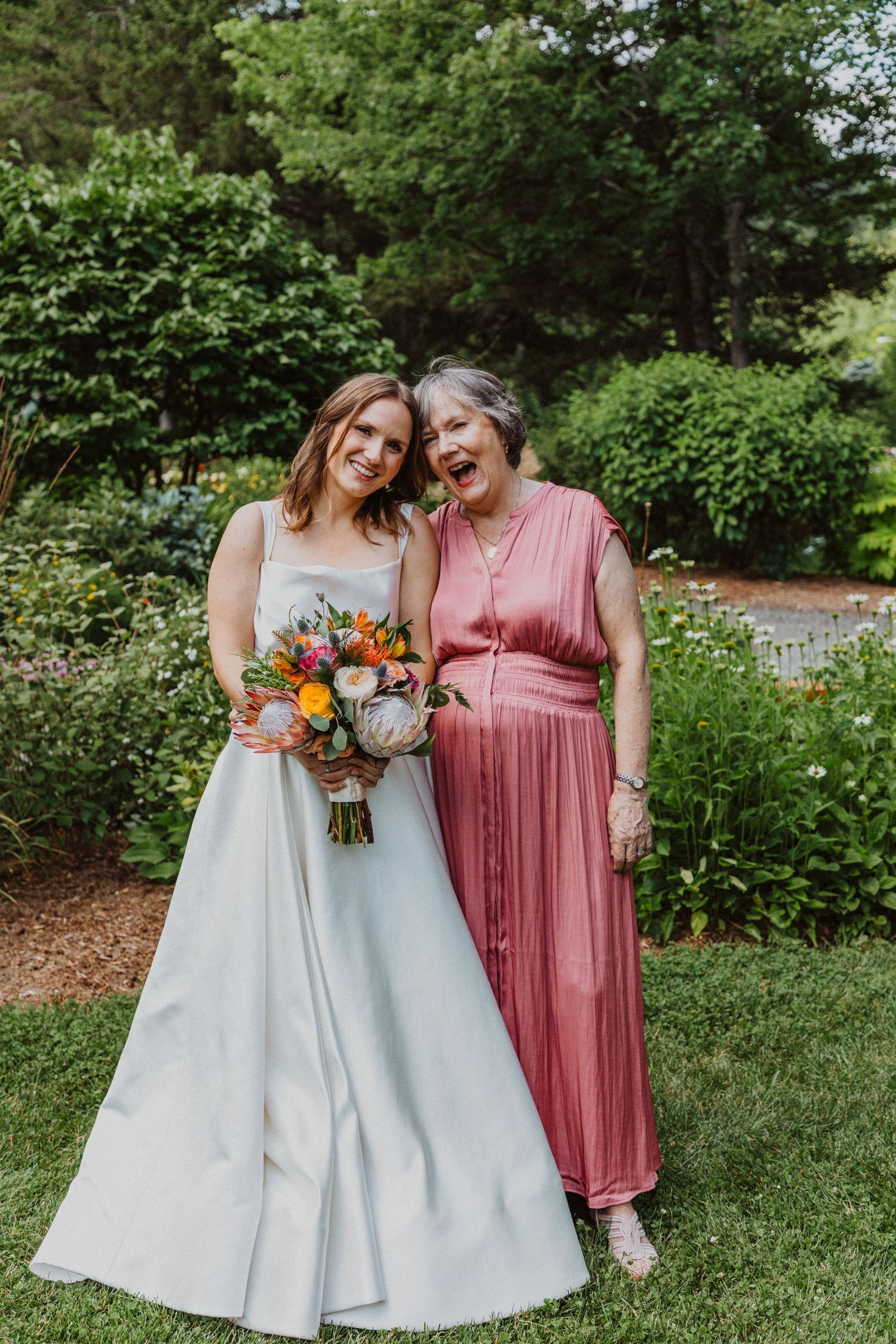 Smiling bride in white gown holding tropical bouquet poses with elderly woman in pink dress in garden.