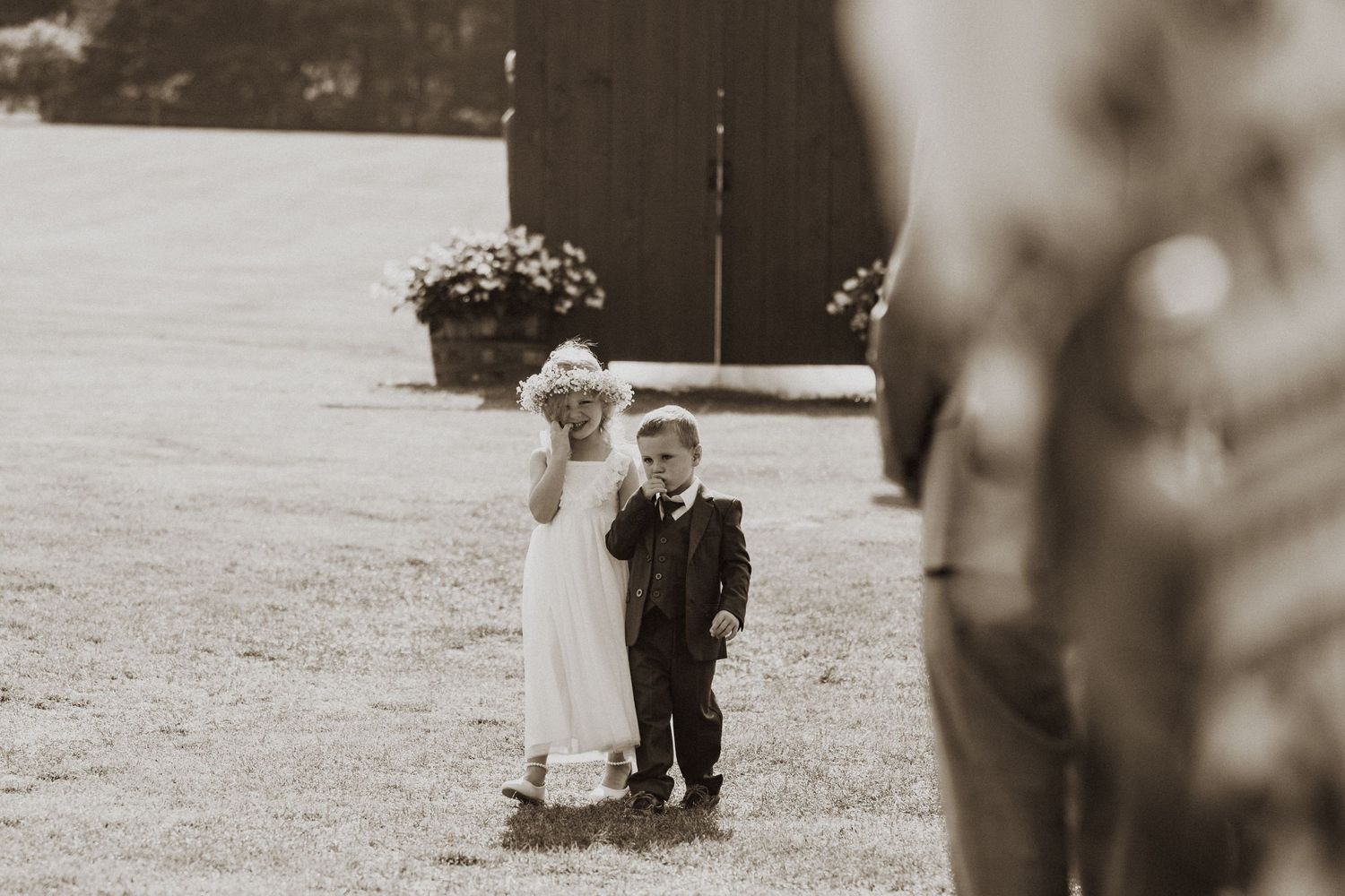 Flower girl and ring bearer walking together outdoors near dark barn in sepia photo.