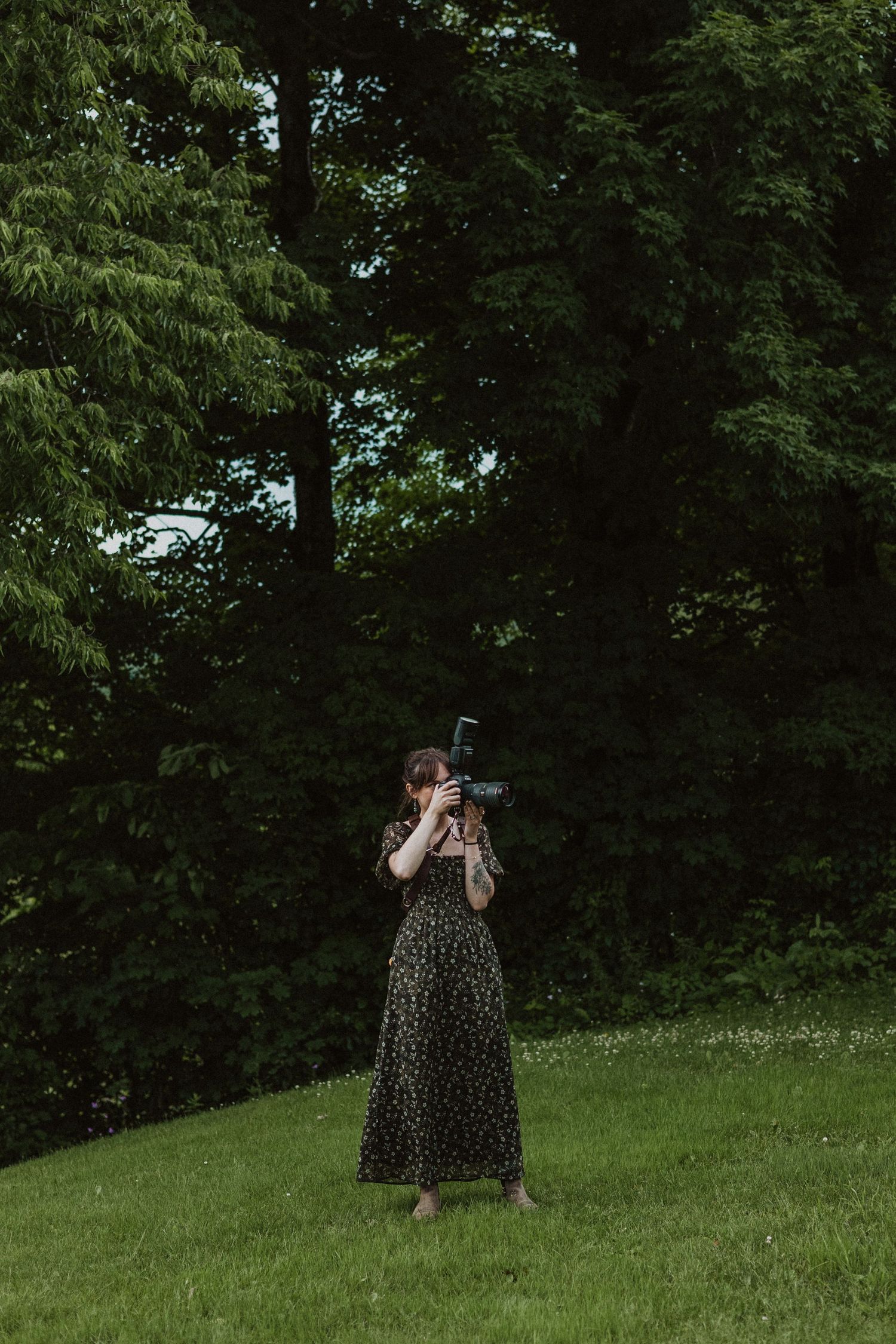 Female photographer in floral dress photographing outdoors in a lush green forest setting.