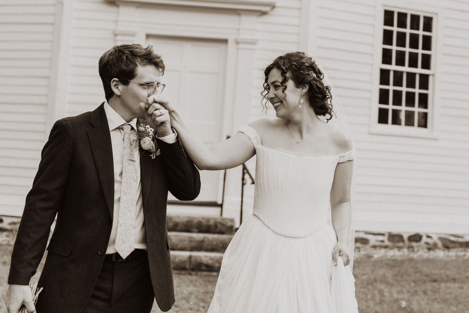 Groom kissing bride's hand while she laughs in front of a white chapel in a romantic sepia-toned wedding photo.