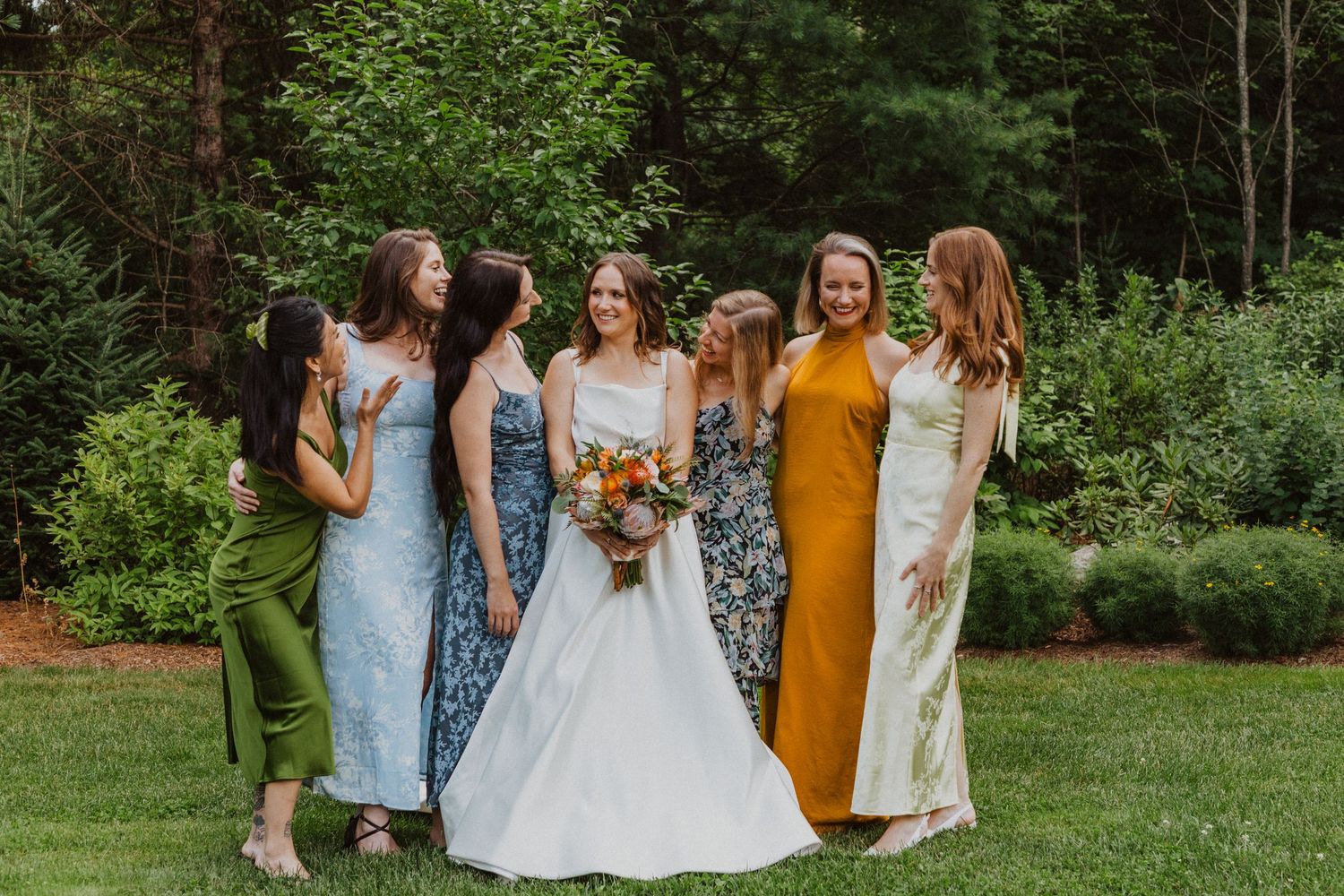 Bride in white gown poses with six bridesmaids in colorful dresses outdoors in a lush green garden setting.