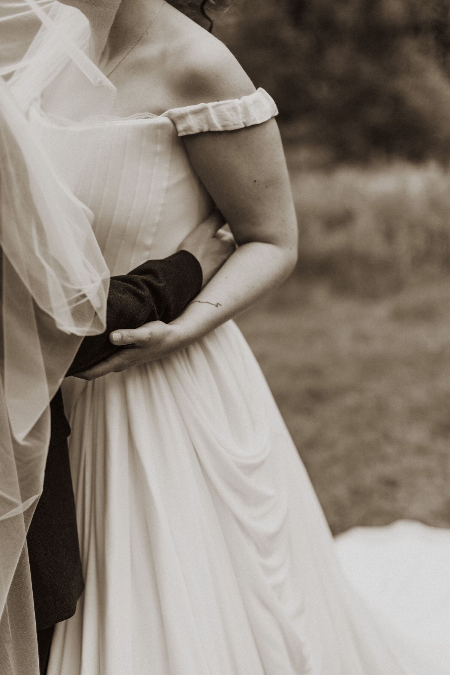 Sepia close-up of groom's hands tenderly embracing bride's waist in flowing off-shoulder gown.