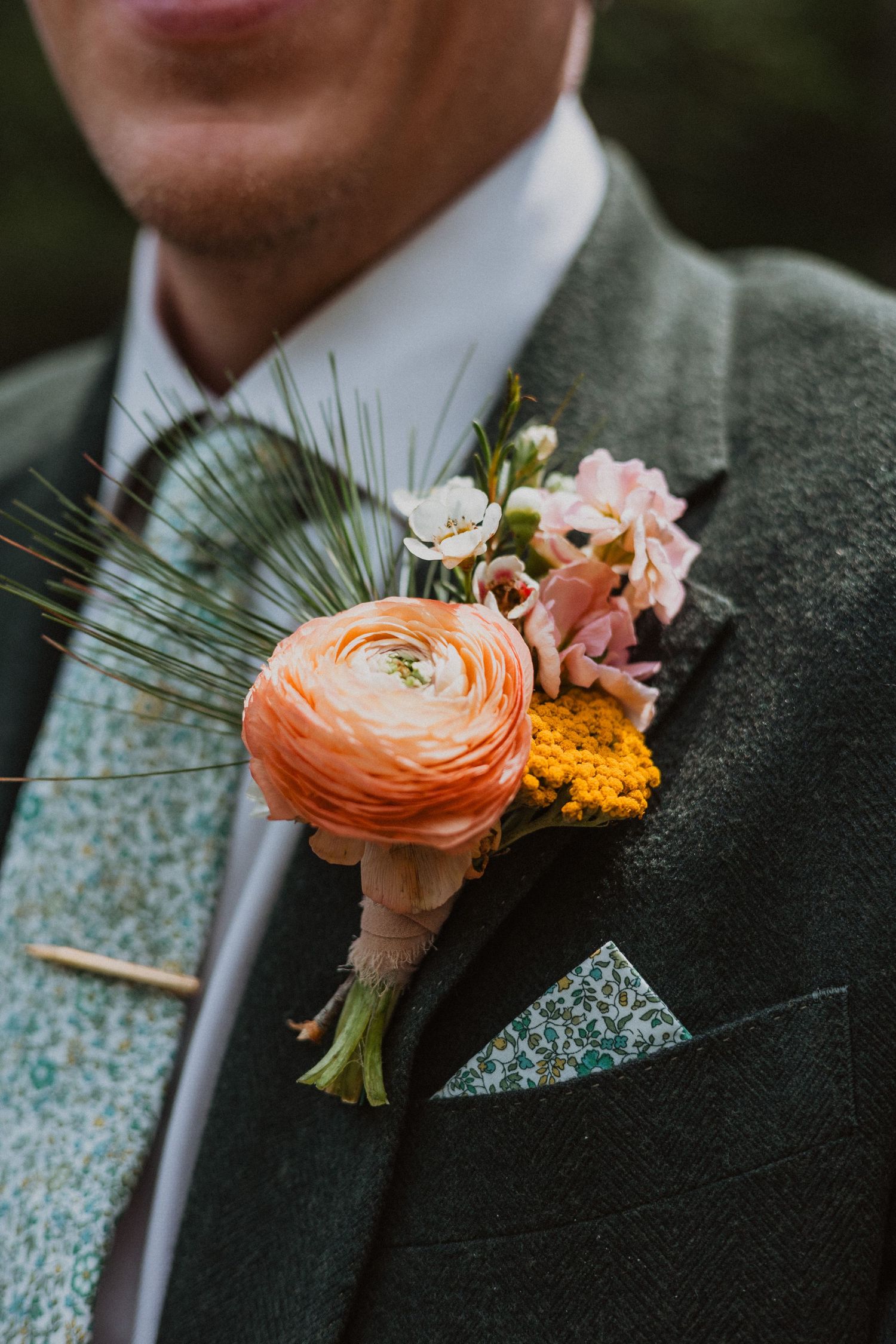 Groom wearing green tweed suit with orange ranunculus boutonniere featuring pink flowers and yellow accents.