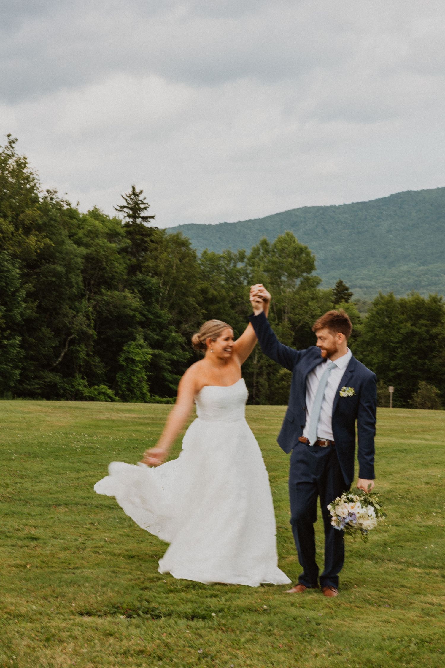 Groom twirling bride in a white strapless gown on a green field with mountain backdrop.