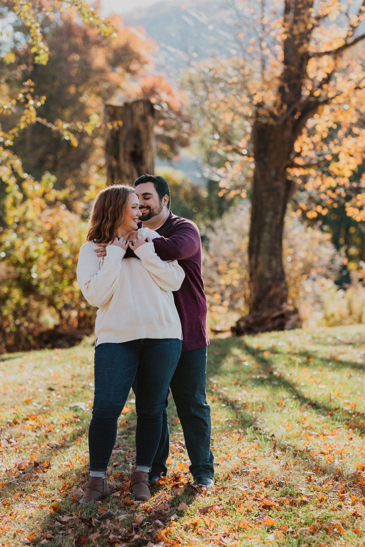 Couple embracing in a sunlit autumn park, surrounded by golden fall foliage and colorful leaves on the ground.