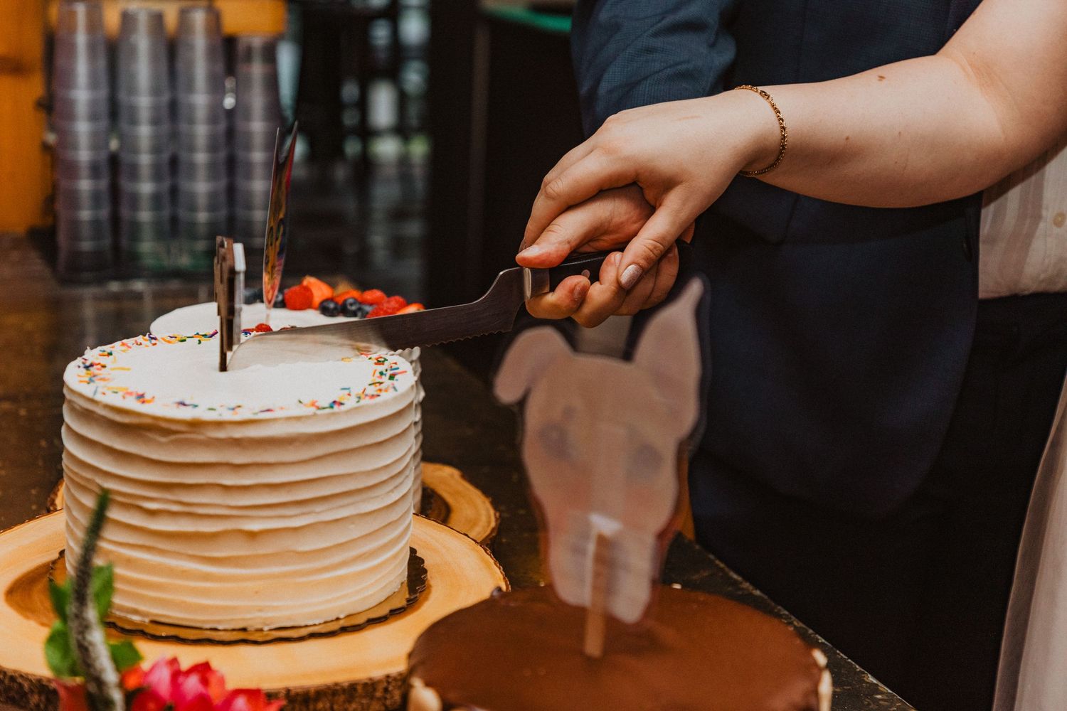 Bride and groom cut elegant white layered wedding cake displayed on a rustic wood slice at their reception.