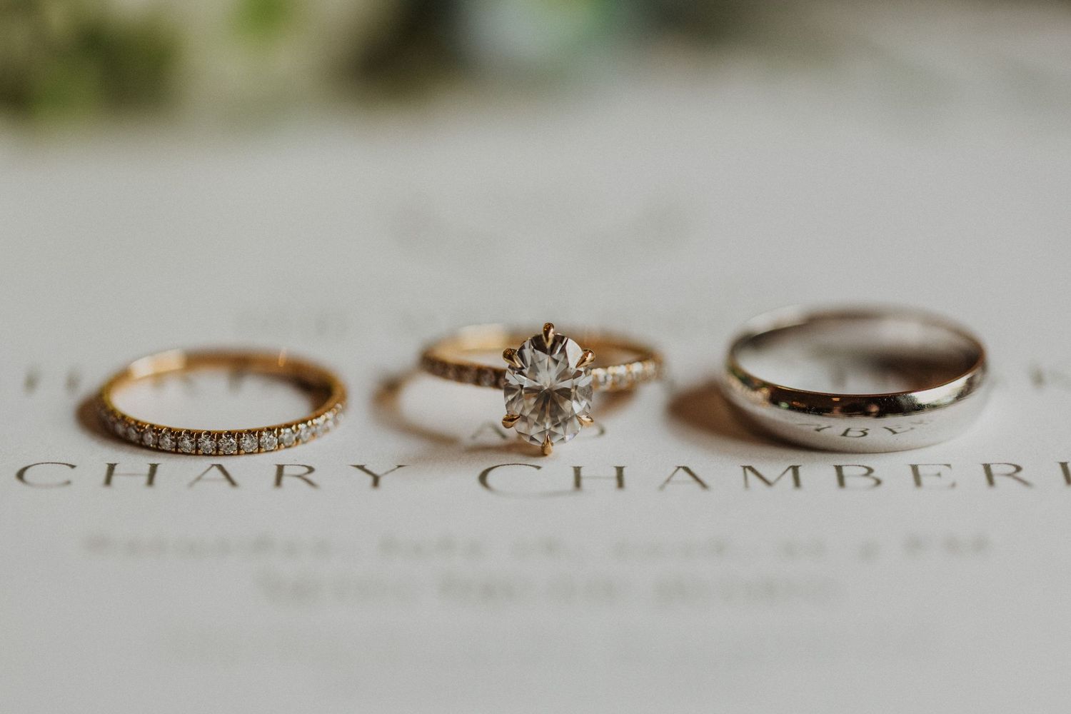 Three elegant wedding rings displayed together, featuring a diamond engagement ring flanked by gold and silver bands on white paper.
