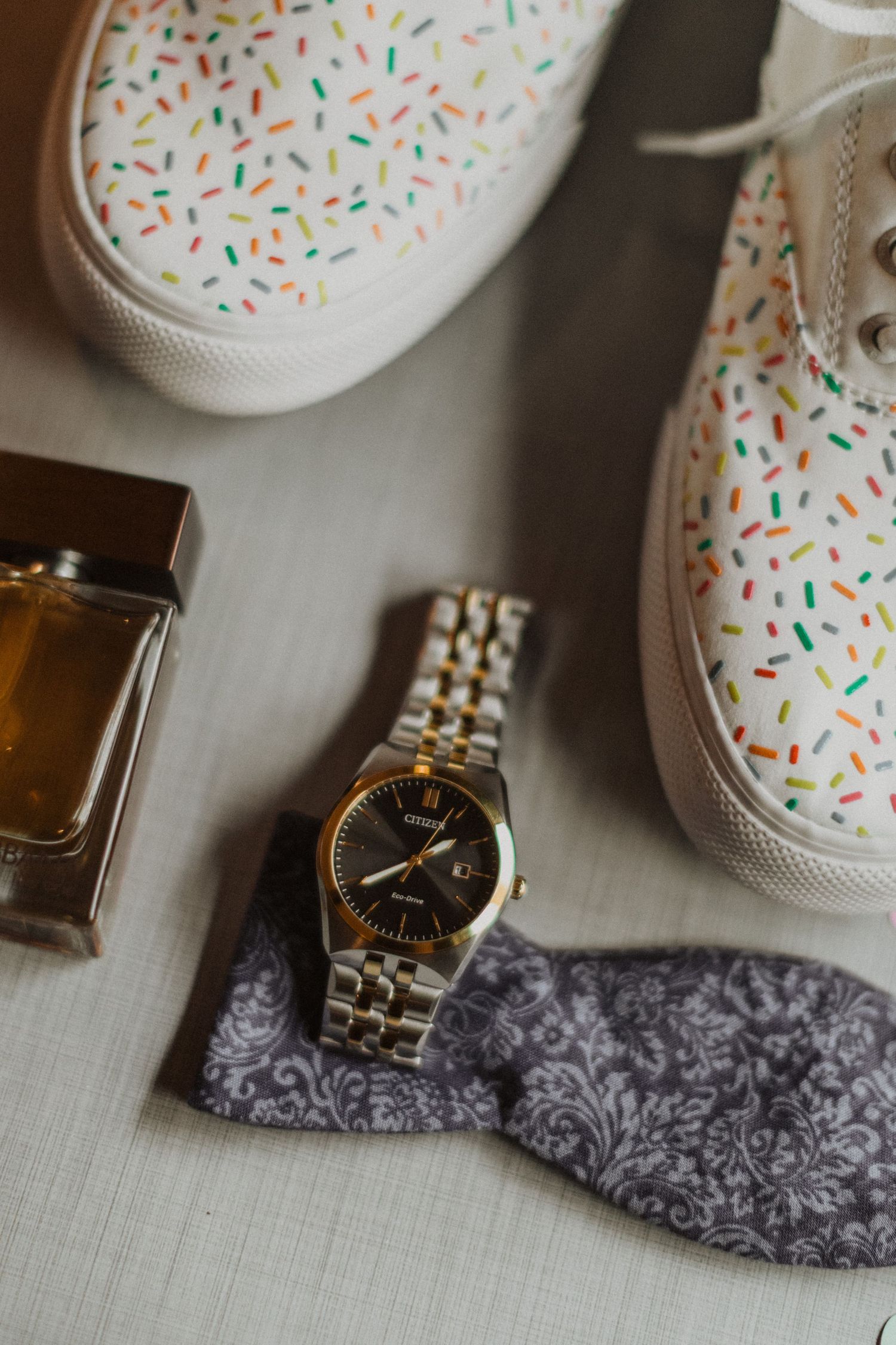 Groom wedding accessories flat lay featuring sprinkle print sneakers, gold watch, cologne, and gray patterned pocket square.
