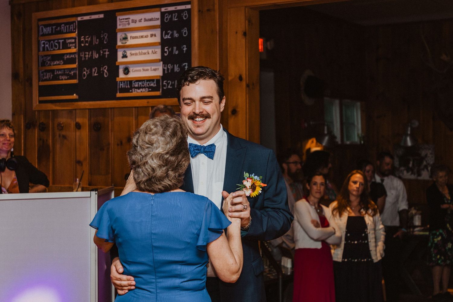 Groom in navy suit and bow tie sharing mother-son dance at rustic barn wedding reception.
