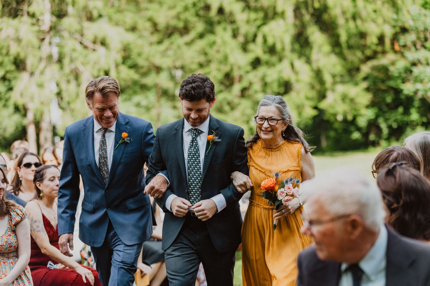Groom walks down outdoor wedding aisle escorted by two family members, surrounded by seated guests among lush green trees.
