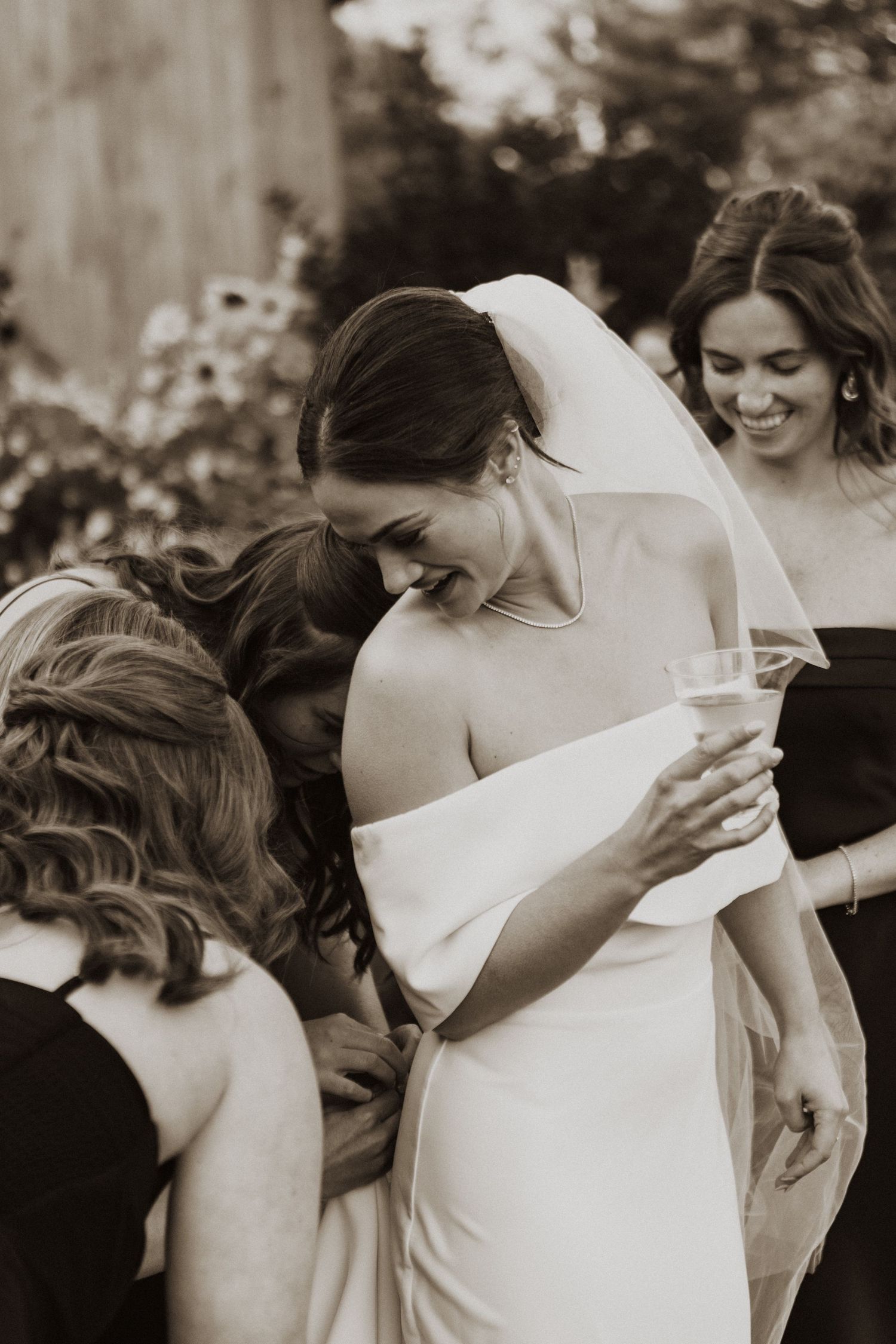 Black and white photo of a smiling bride holding a champagne glass surrounded by guests at her wedding.