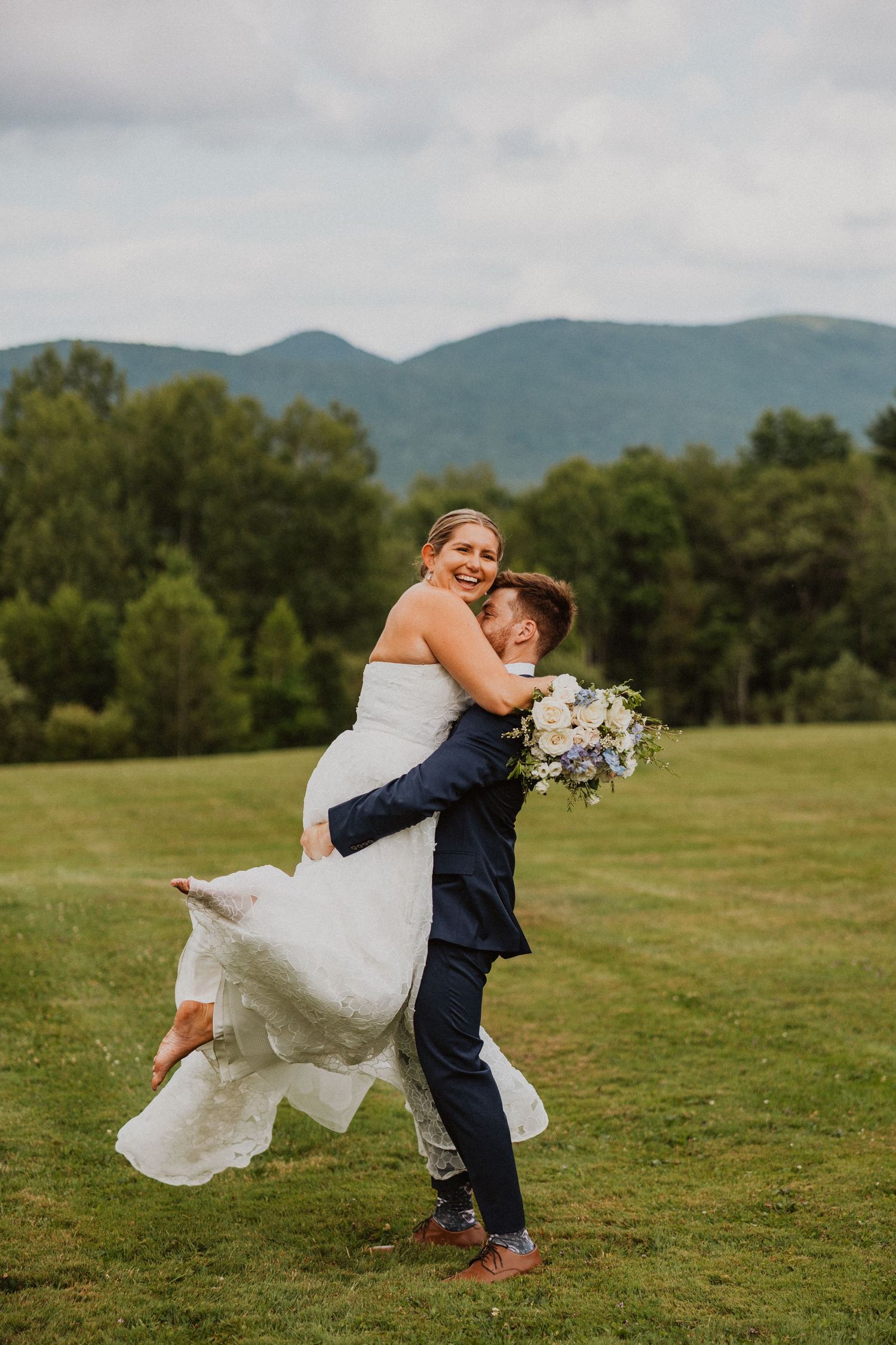 Groom joyfully lifting smiling bride in white wedding gown on a green field with mountains.