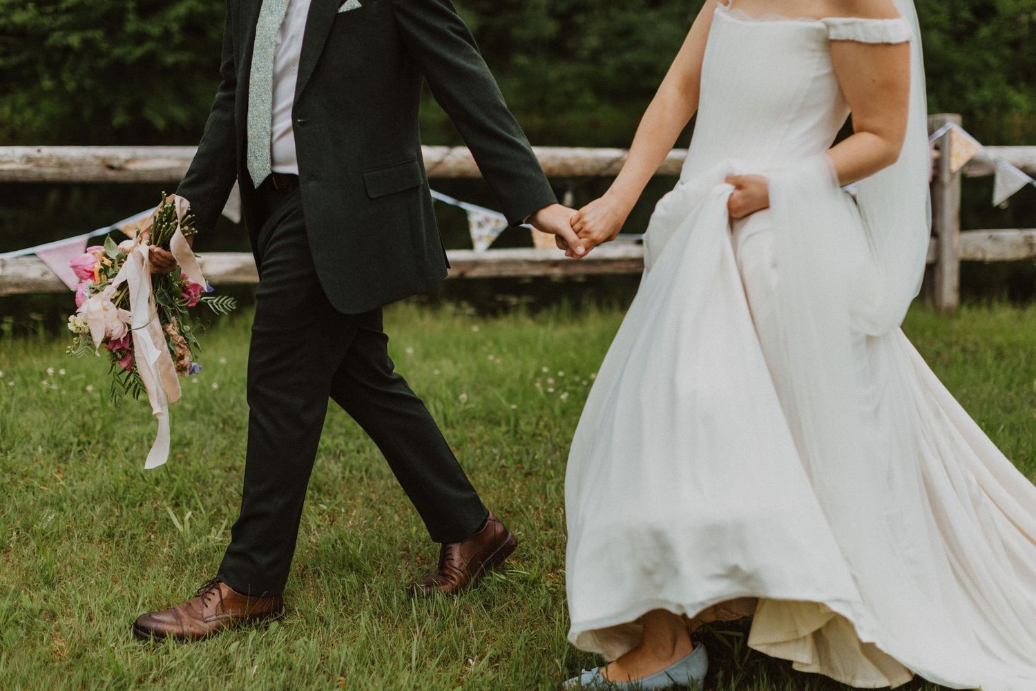 Bride and groom walking hand in hand on green grass, bride in white gown, groom in black suit holding bouquet.