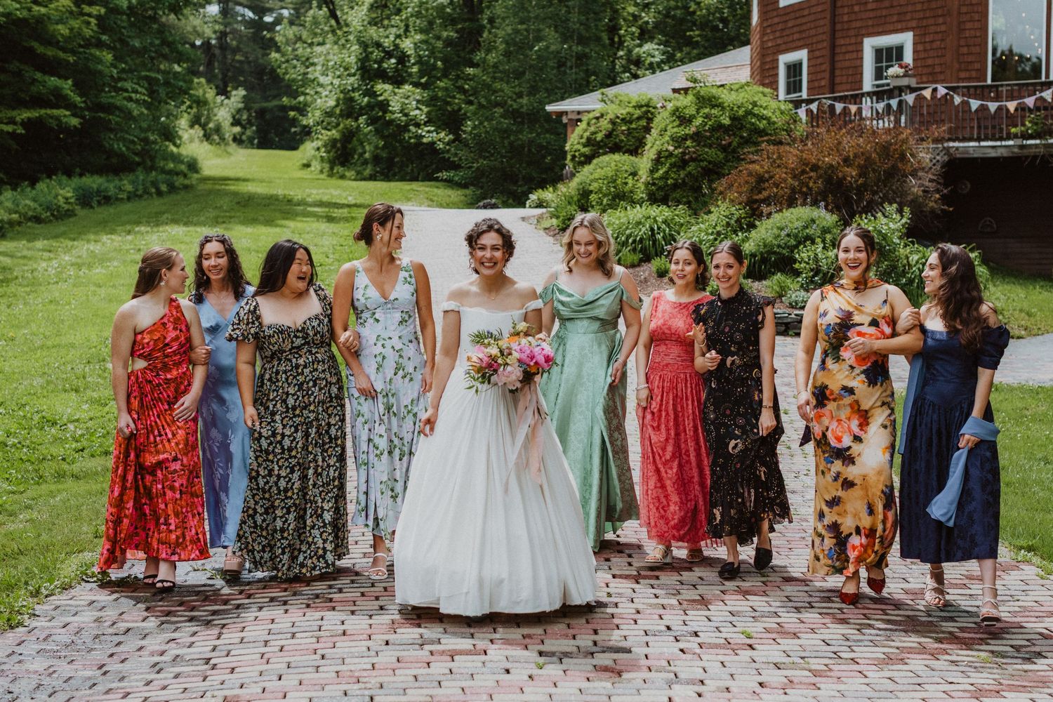 Bride in white ball gown surrounded by bridesmaids in colorful floral dresses on a brick pathway outdoors.