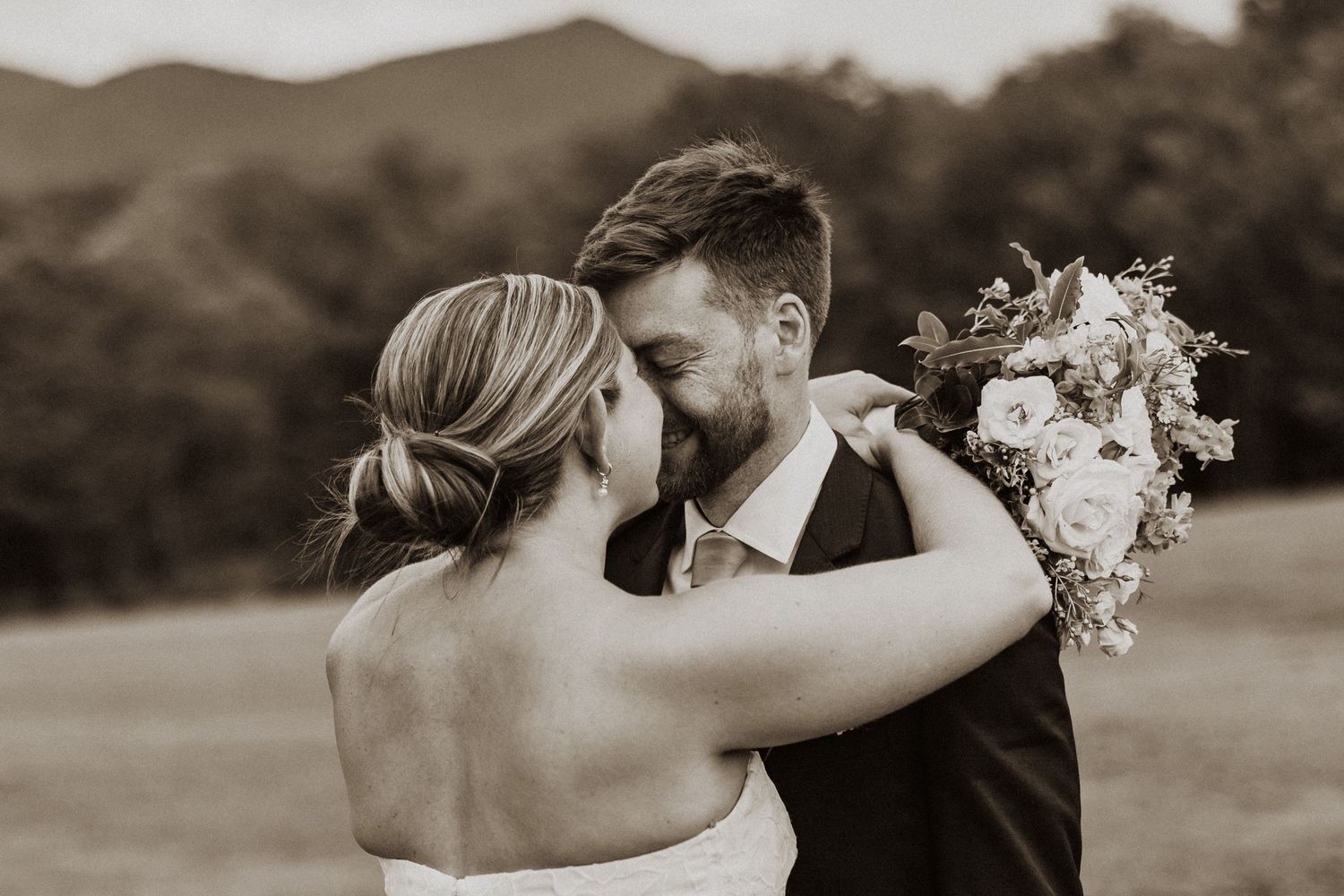 Bride and groom sharing romantic kiss outdoors with mountain views and bridal bouquet in sepia tone.