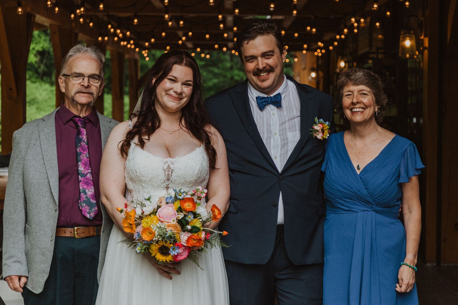 Wedding party of four posing outdoors under string lights, bride holding colorful bouquet with sunflowers.