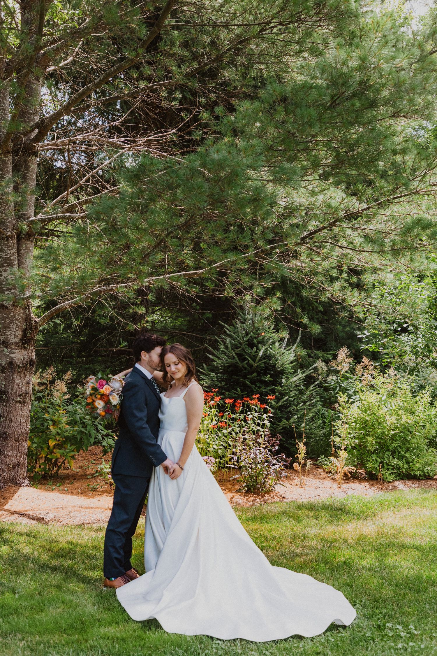 Bride and groom sharing intimate kiss under tall pine tree surrounded by colorful garden on wedding day.