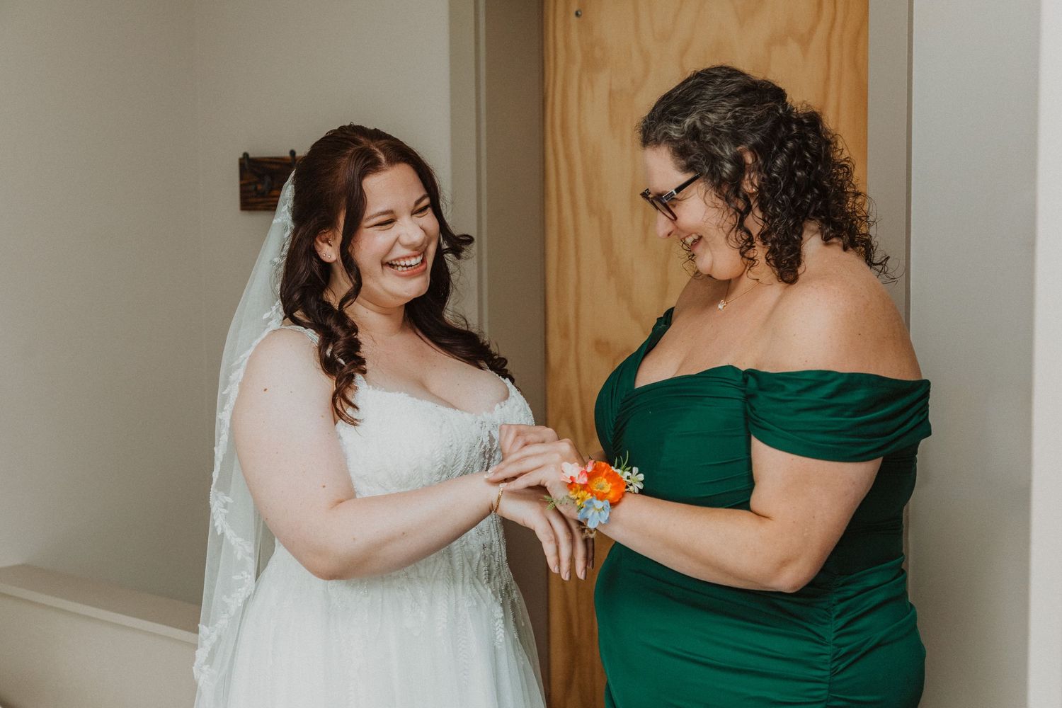 Bride in white gown and veil smiles as bridesmaid in green off-shoulder dress admires orange wrist corsage.