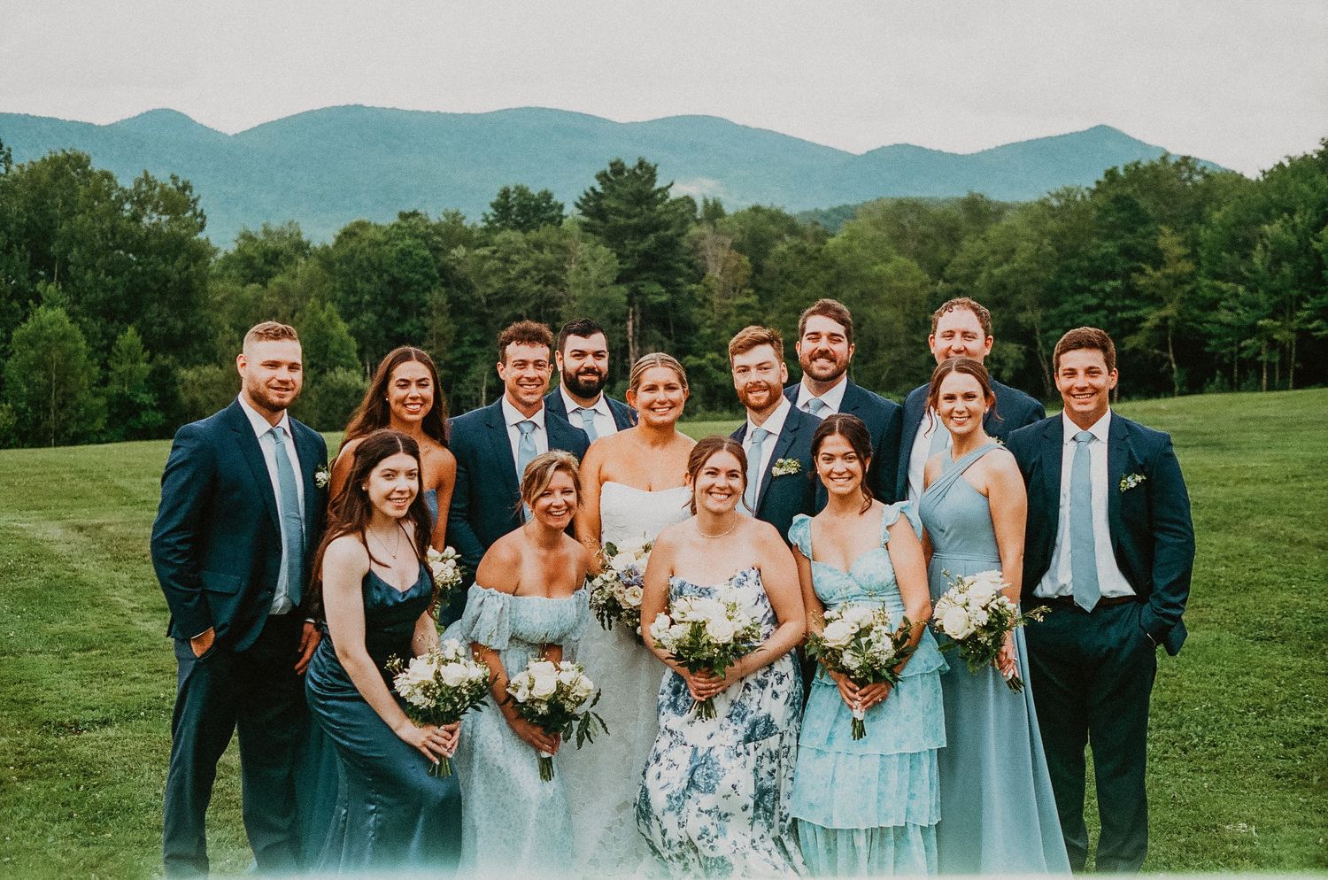 Wedding party poses outdoors with mountain backdrop, bridesmaids in blue dresses and groomsmen in navy suits.