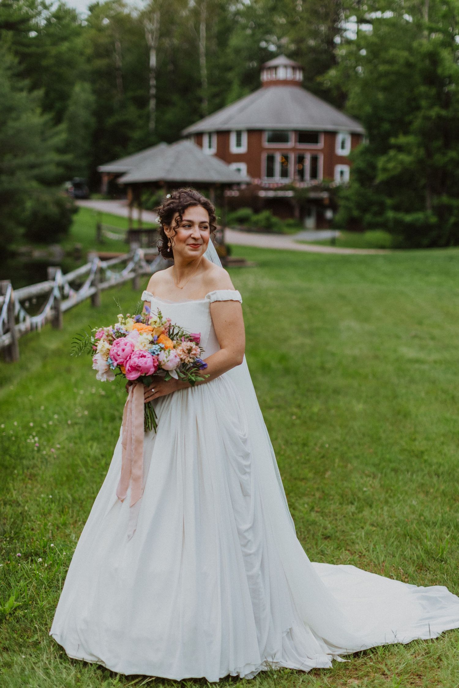Bride in off-shoulder white gown holding colorful bouquet stands on green lawn with rustic red building backdrop.