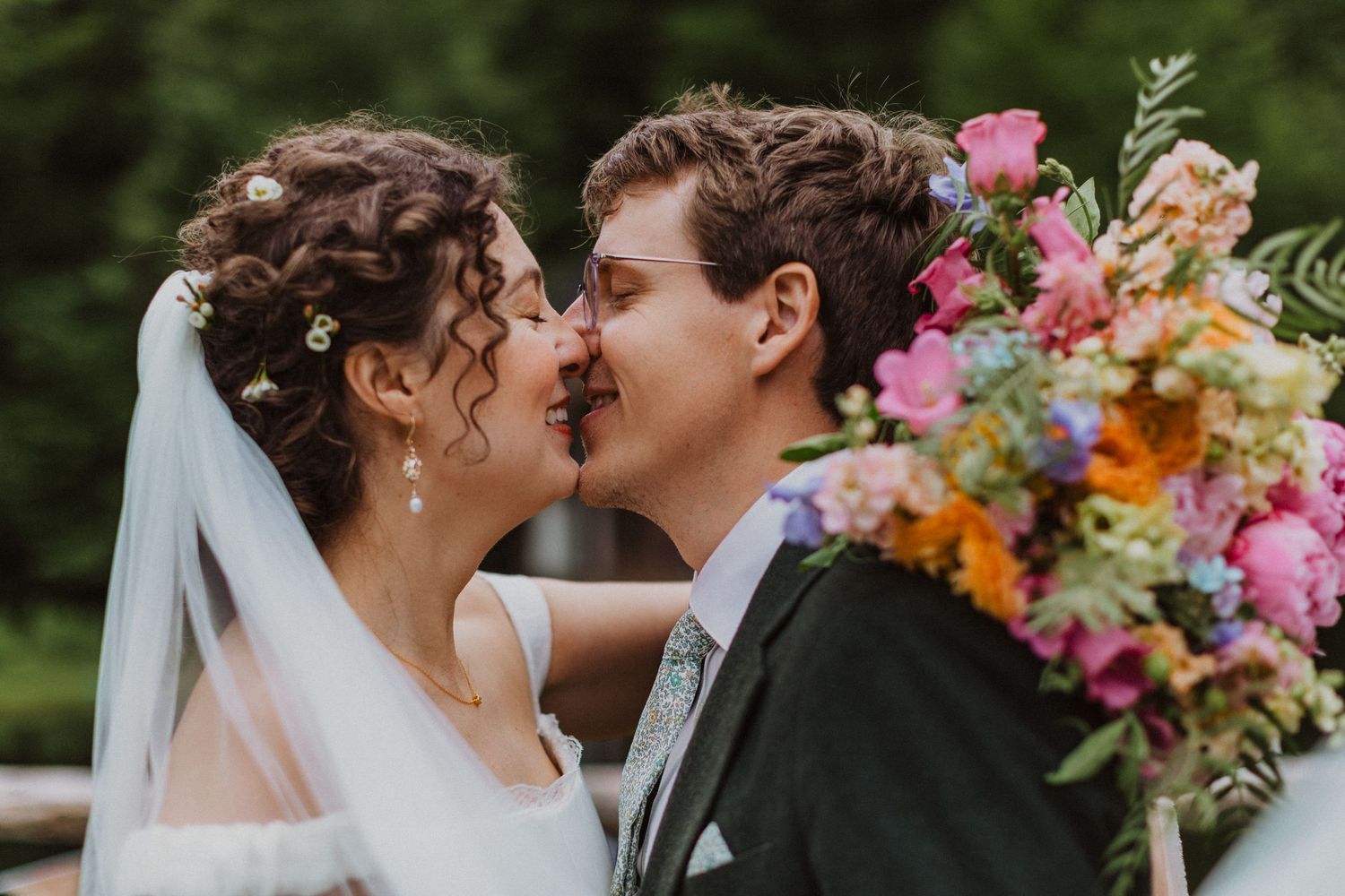 Bride and groom sharing a romantic kiss surrounded by a vibrant colorful wedding bouquet outdoors.
