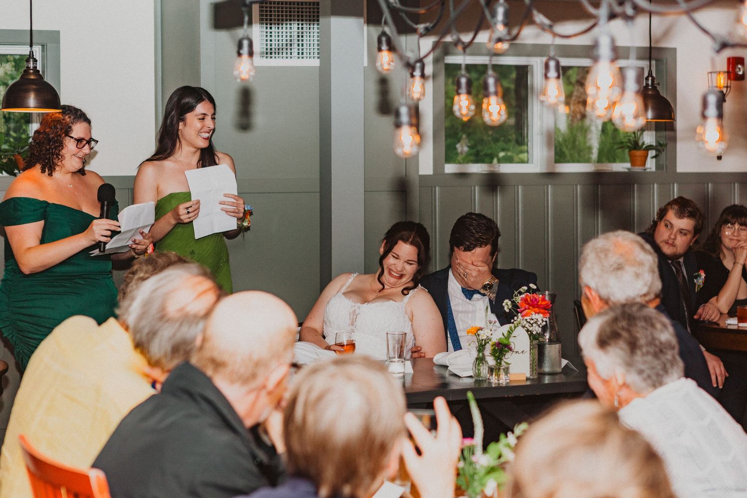 Wedding reception toast with bridesmaid in green dress reading speech while bride and groom smile at decorated table.