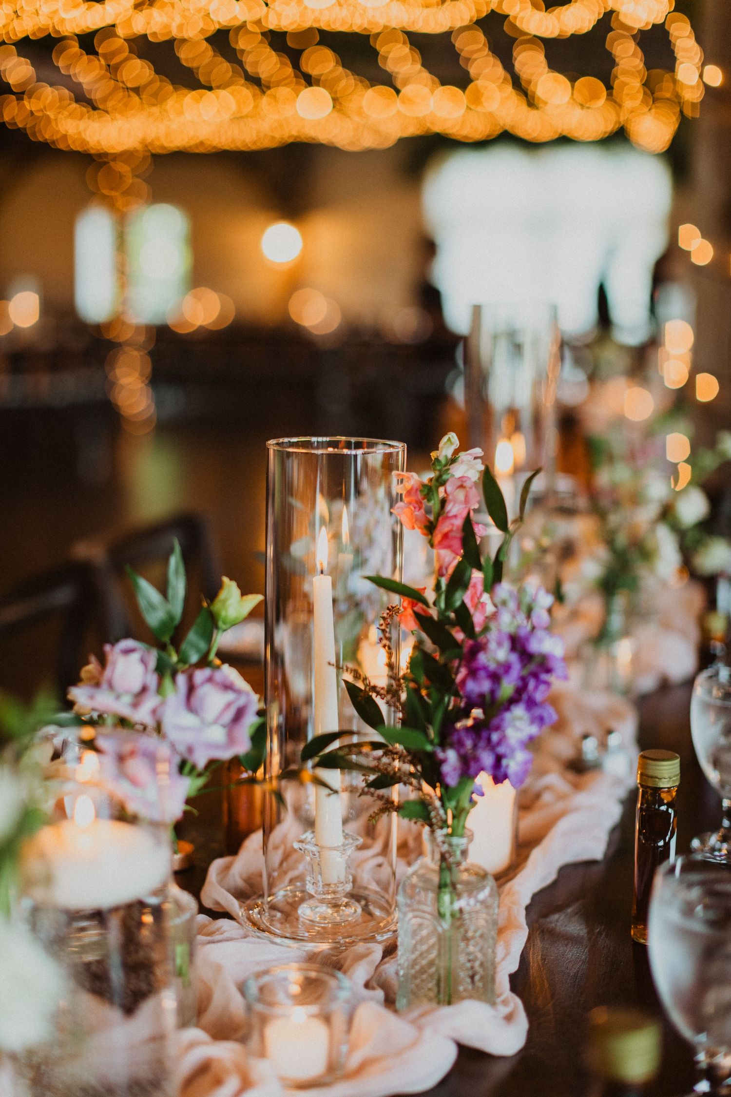 Elegant wedding reception table with candles, colorful florals, and warm string lights overhead.