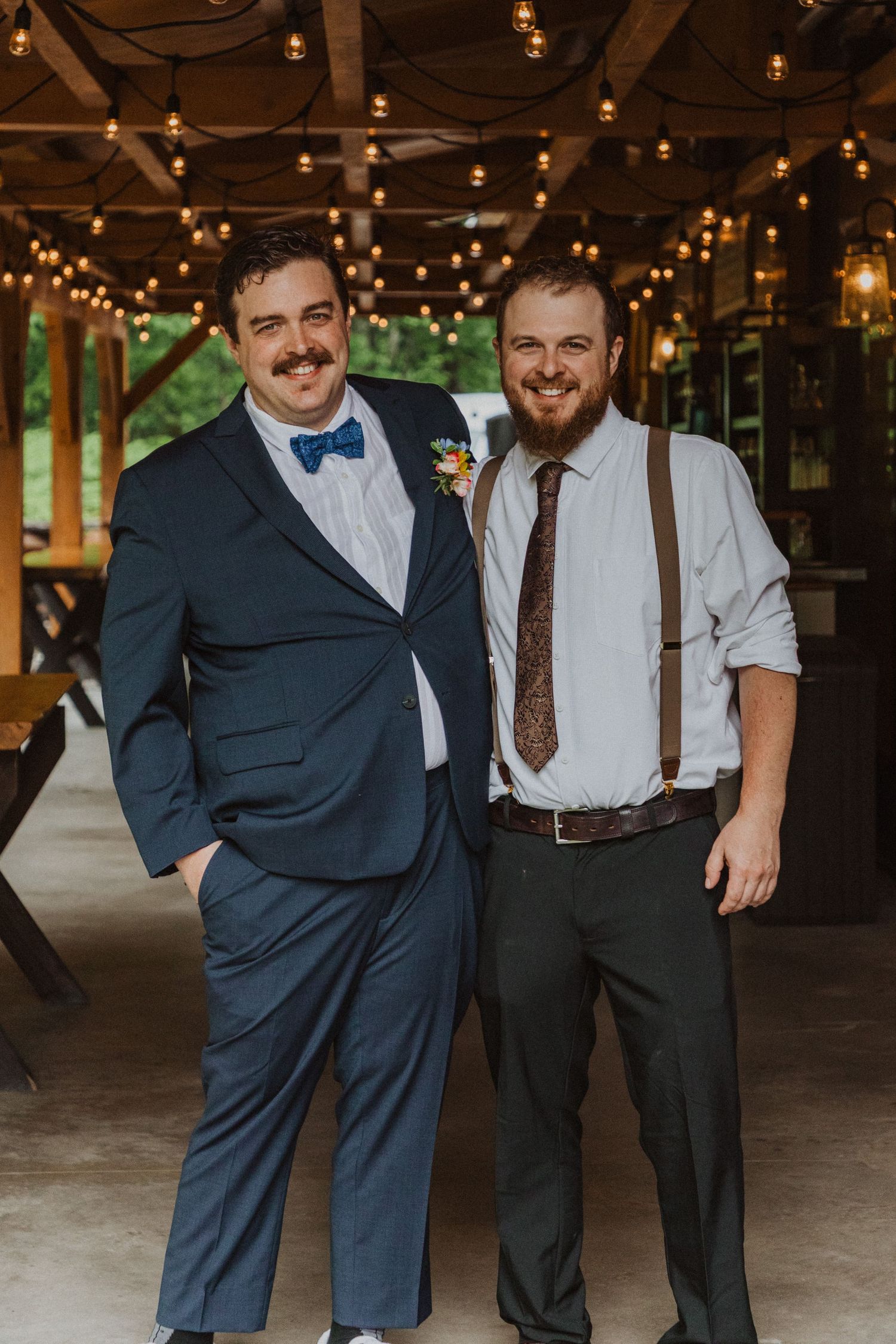 Two smiling men in formal attire pose together at a rustic wedding venue with warm string lights overhead.