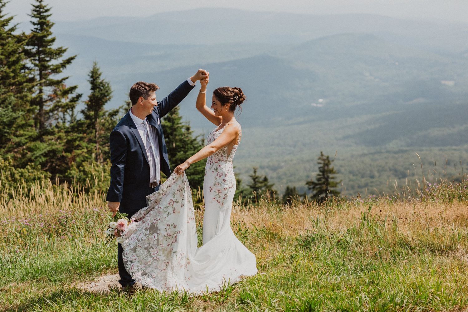 Bride and groom dancing on mountaintop meadow, bride in floral gown with scenic mountain valley backdrop.