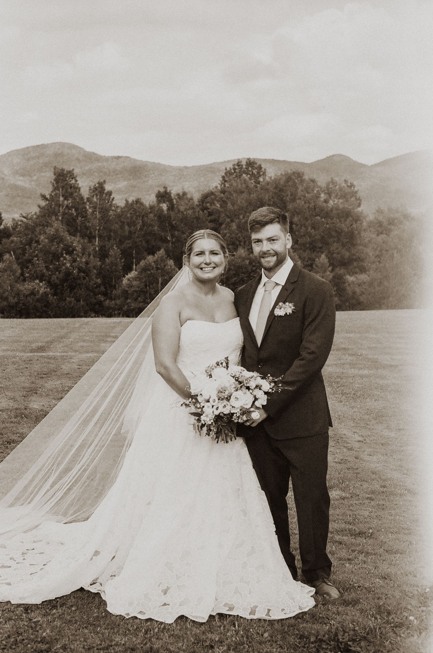 Vintage sepia portrait of bride and groom posing with bouquet on a scenic mountain field.