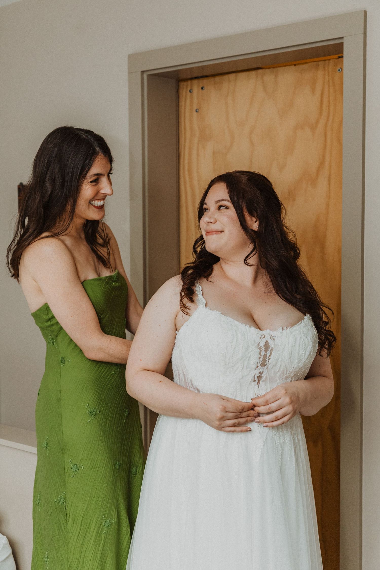 Bridesmaid in green dress helping bride in white wedding gown get ready before the ceremony.