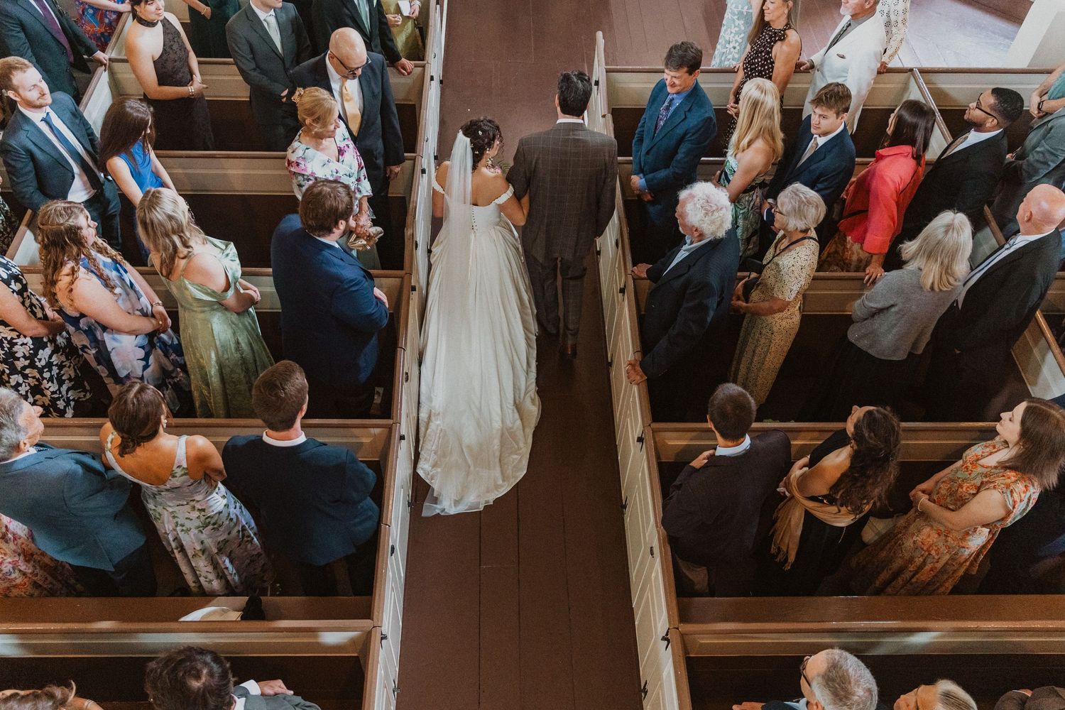 Aerial view of bride walking down church aisle lined with seated wedding guests in wooden pews.