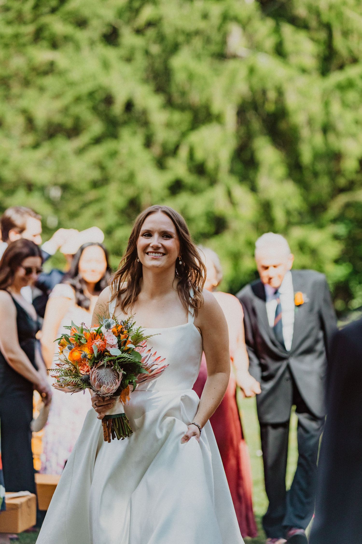 Radiant bride walking down outdoor aisle in white gown holding vibrant orange and pink tropical bouquet.