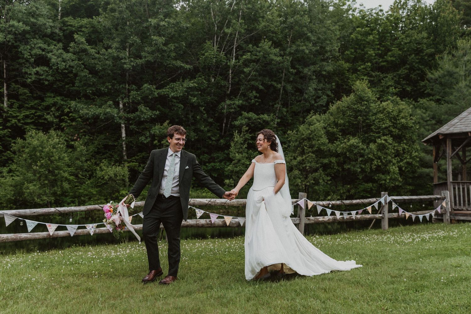 A bride and groom hold hands walking across a green lawn with a rustic fence and forest backdrop.