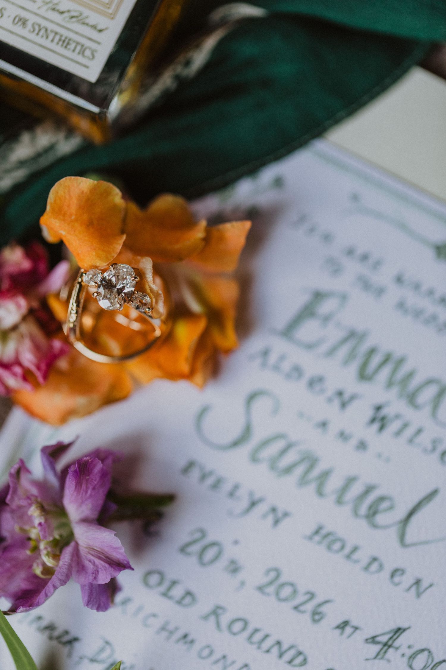 Diamond engagement ring resting on colorful orange and purple flowers beside elegant wedding invitation calligraphy.