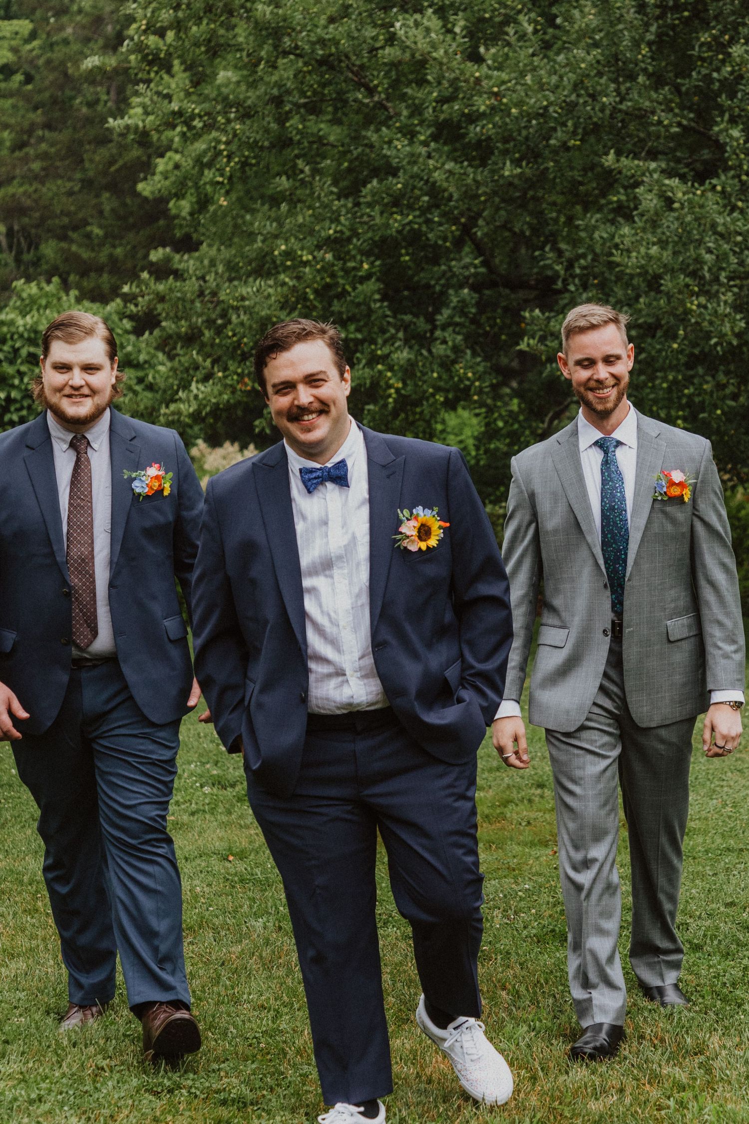 Three groomsmen in navy and gray suits with sunflower boutonnieres walking outdoors on green grass at a wedding.