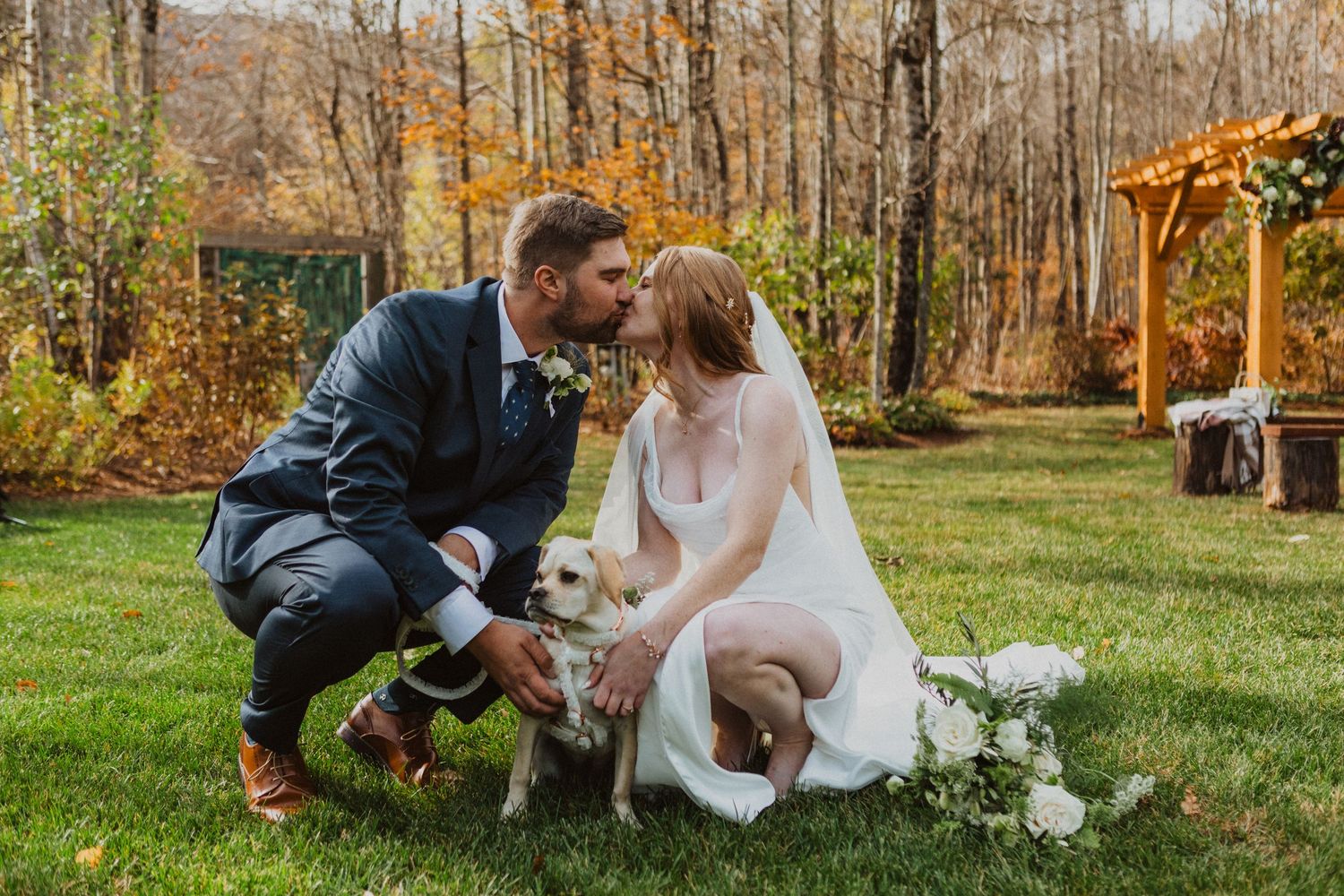 Bride and groom kissing over their white puppy on green grass at an outdoor fall wedding ceremony.
